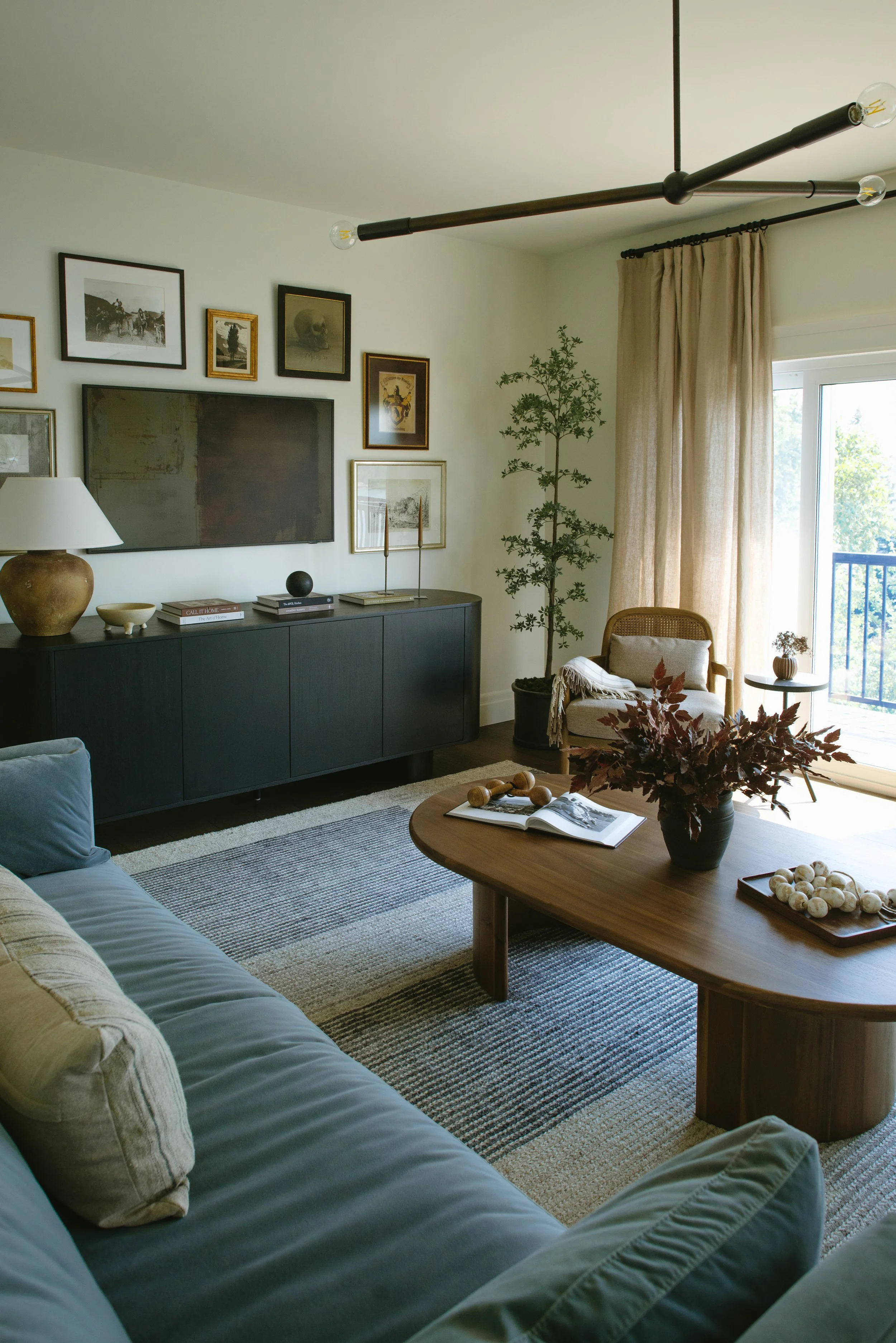 Living room with a dark sideboard, framed art on the wall, a flat-screen TV, a beige armchair near a sliding door, a tall plant, a wooden coffee table with decorative objects, a blue sofa with pillows, and beige curtains.