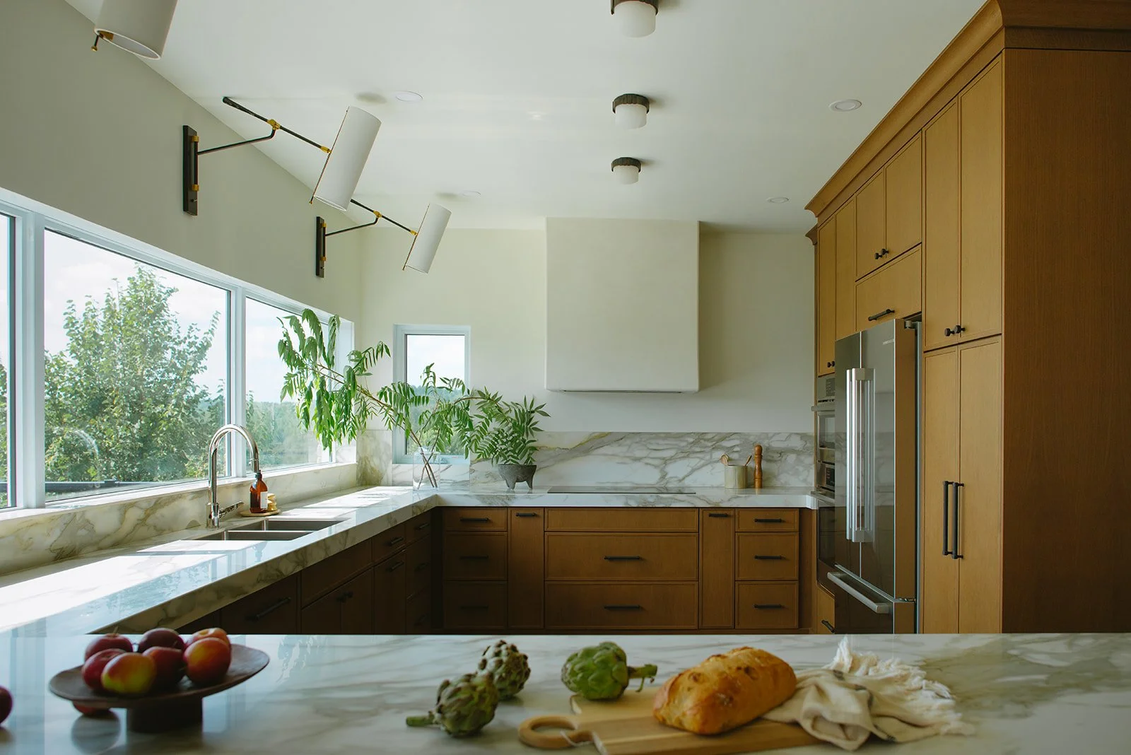 Modern kitchen with marble countertops, wooden cabinets, large window with green plants, and apples, bread, artichokes on the counter.