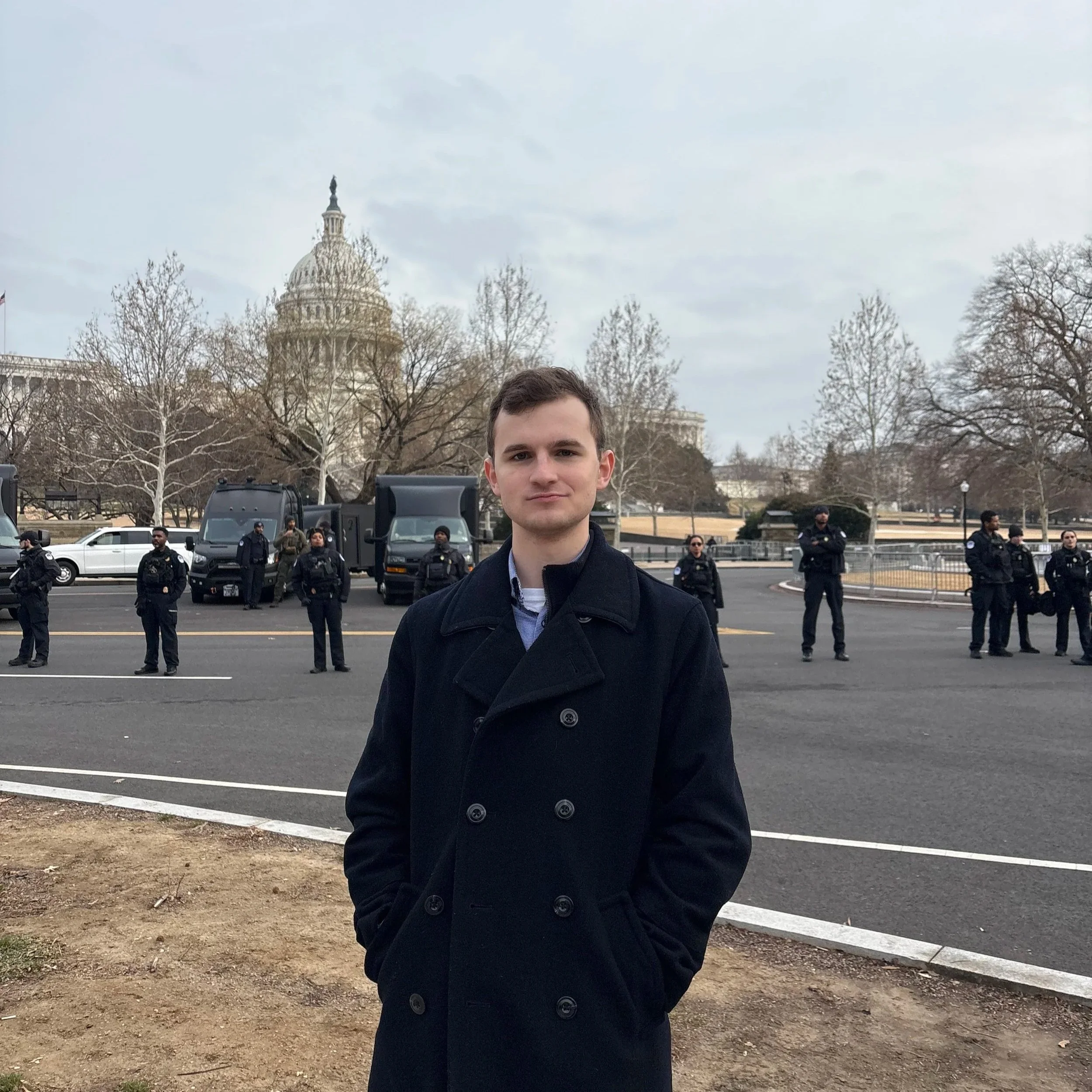 Brady Knox stands in front of a line of police officers and vehicles with the U.S. Capitol building in the background.