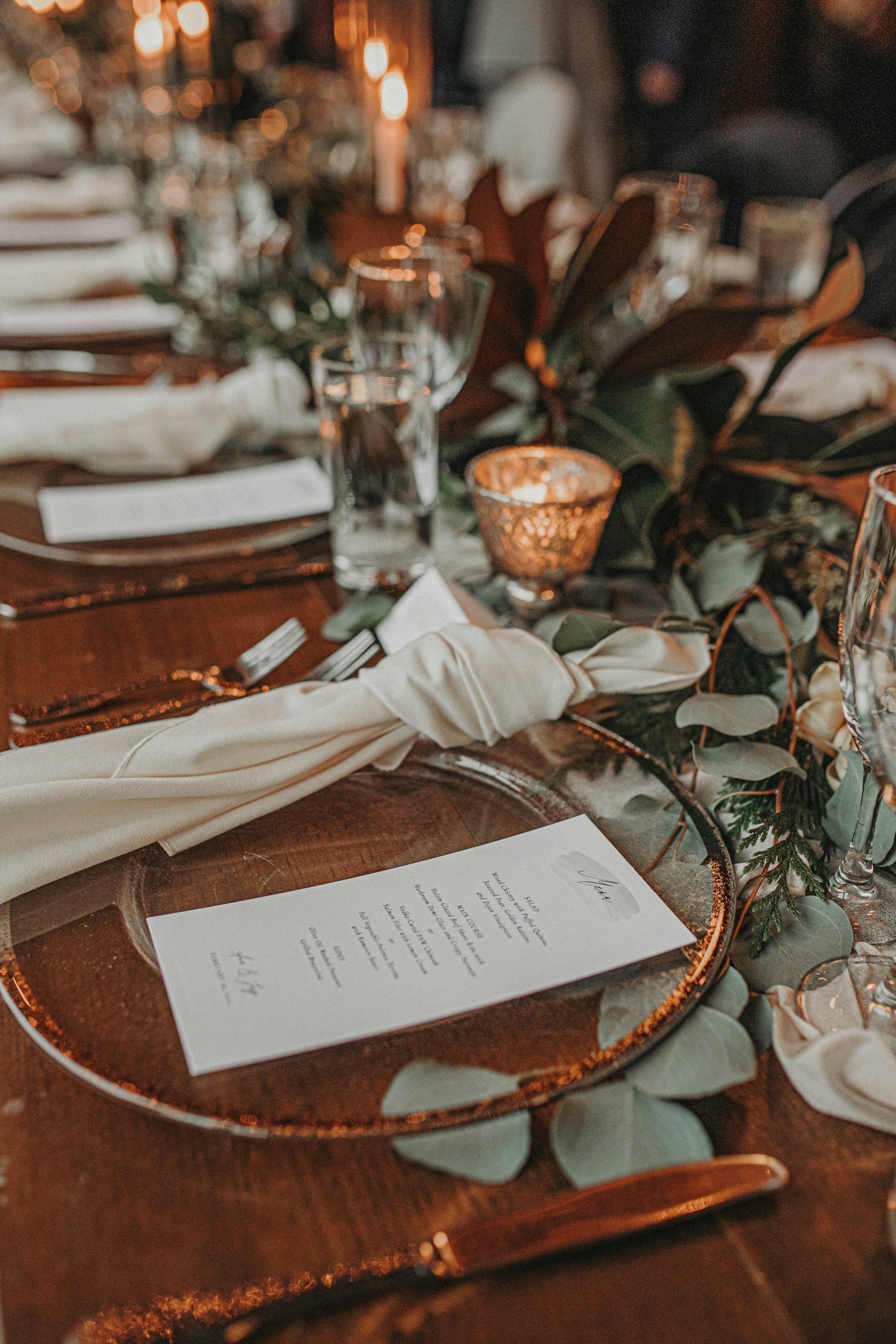 Elegant table setting with a menu card, cream napkin, glassware, and greenery decoration, illuminated by warm candlelight.