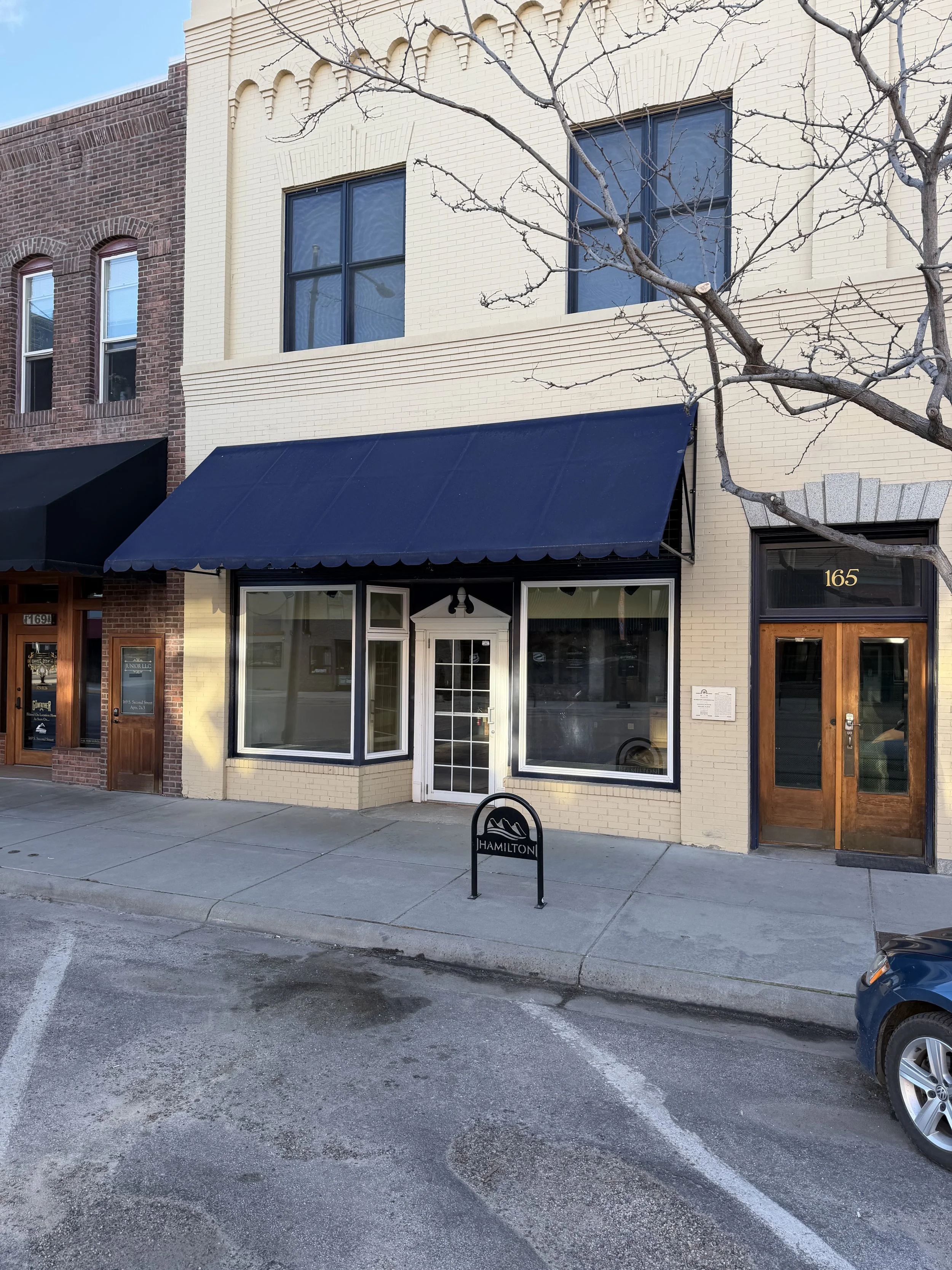 Front view of a building with a black awning, white brick exterior, and large windows. There is a sidewalk and a street with parked cars. A sign reads 'Hamilton.'