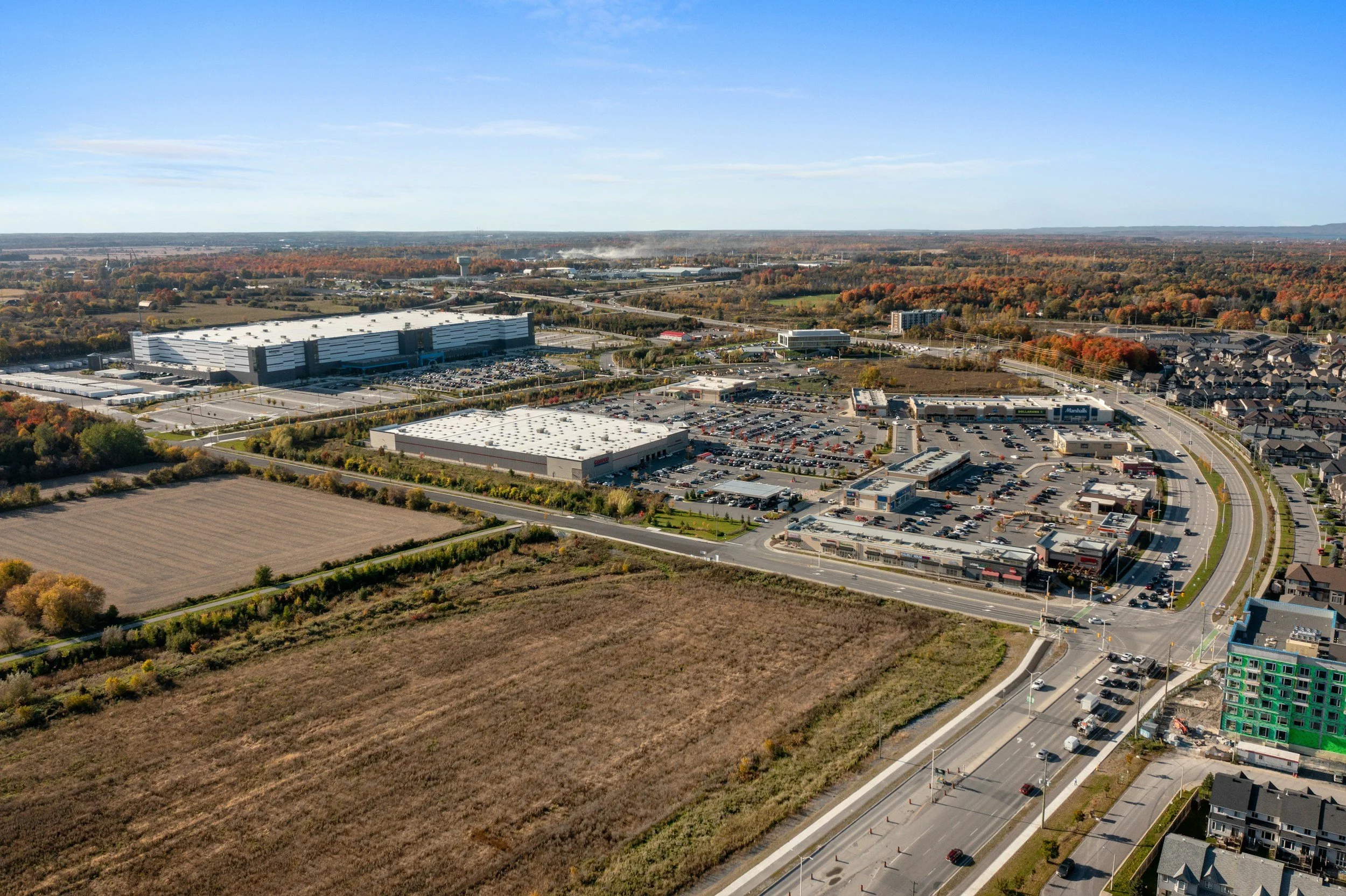 An aerial view of a commercial shopping center with parking lots and surrounding roads, bordered by residential neighborhoods and open fields.