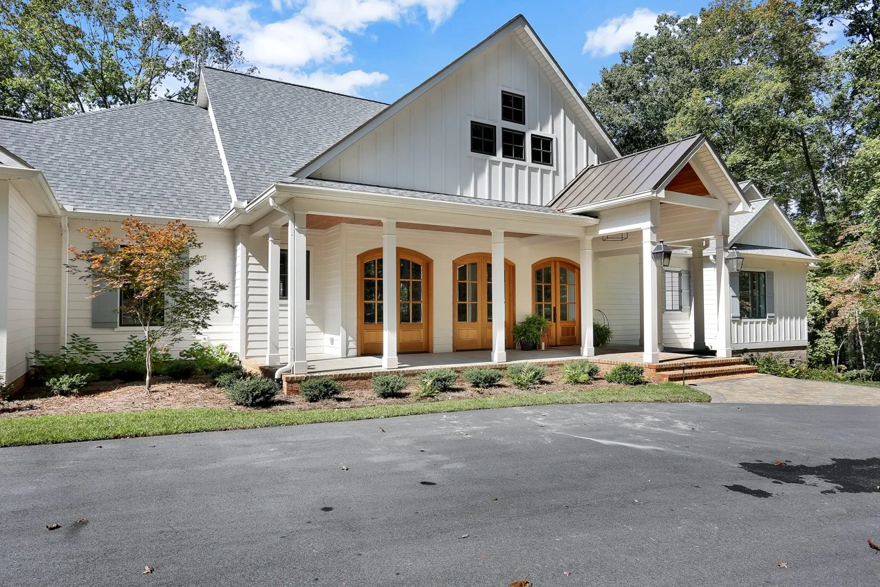 White modern house with front porch, wooden doors, potted plants, and a paved driveway, surrounded by trees under a blue sky.