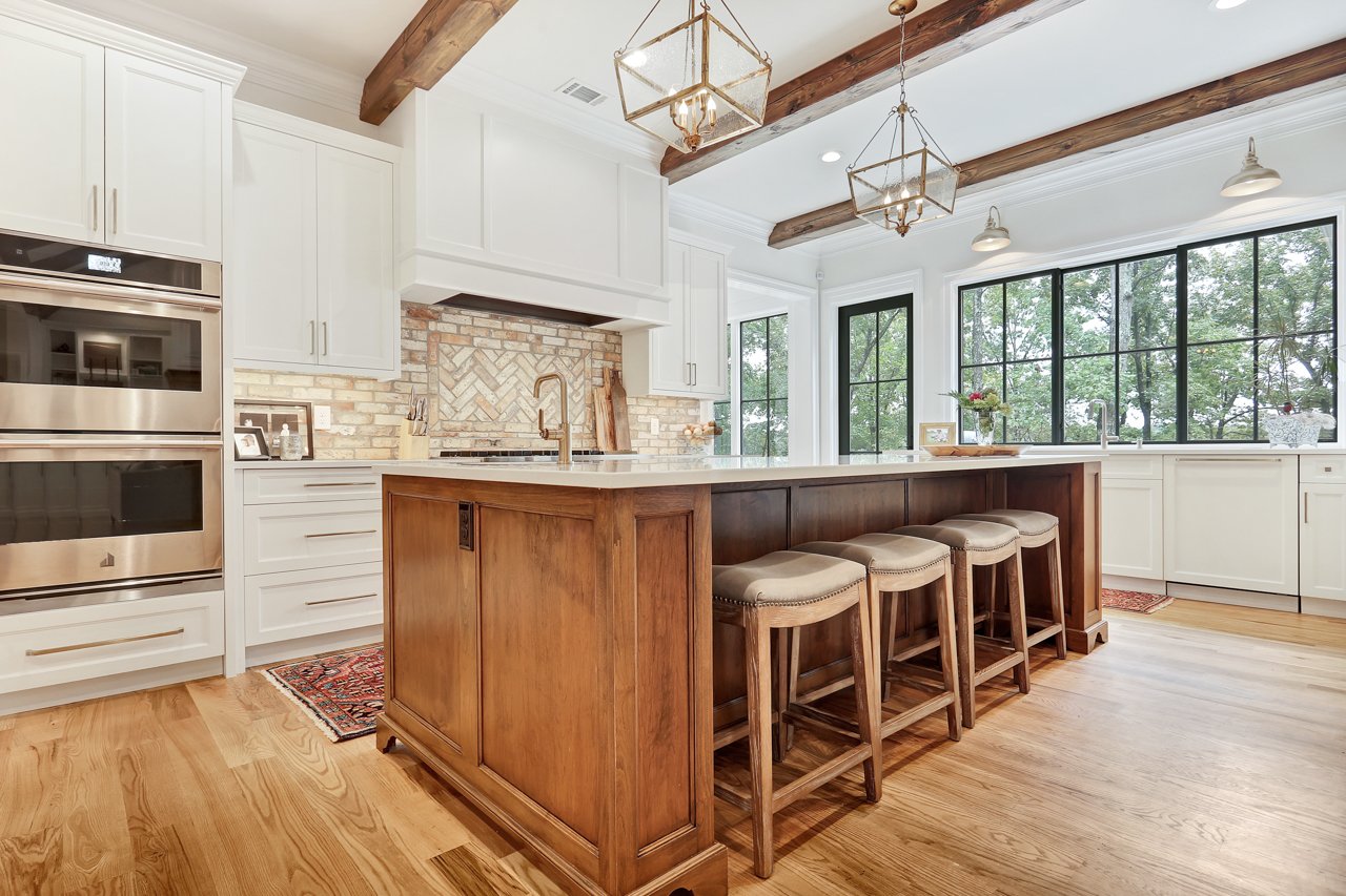 Modern kitchen with white cabinets, wooden island with three stools, brick backsplash, large windows, and wooden ceiling beams.