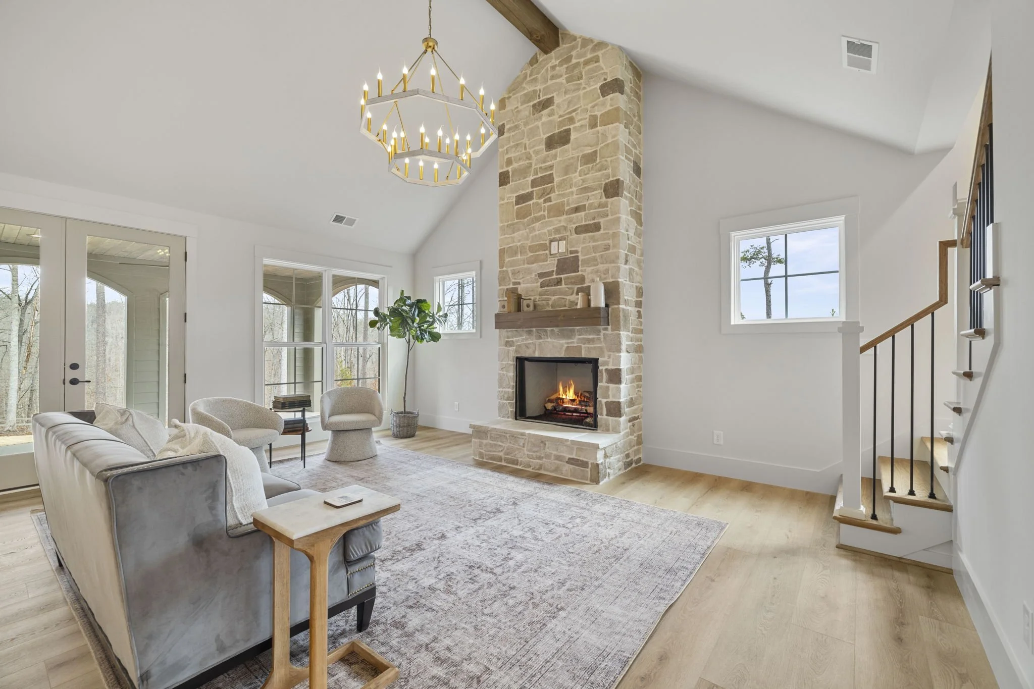 Living room with white walls, wooden flooring, a stone fireplace with a fire, large windows, and a chandelier hanging from the ceiling. There are sofas, chairs, a small table, and a potted plant.