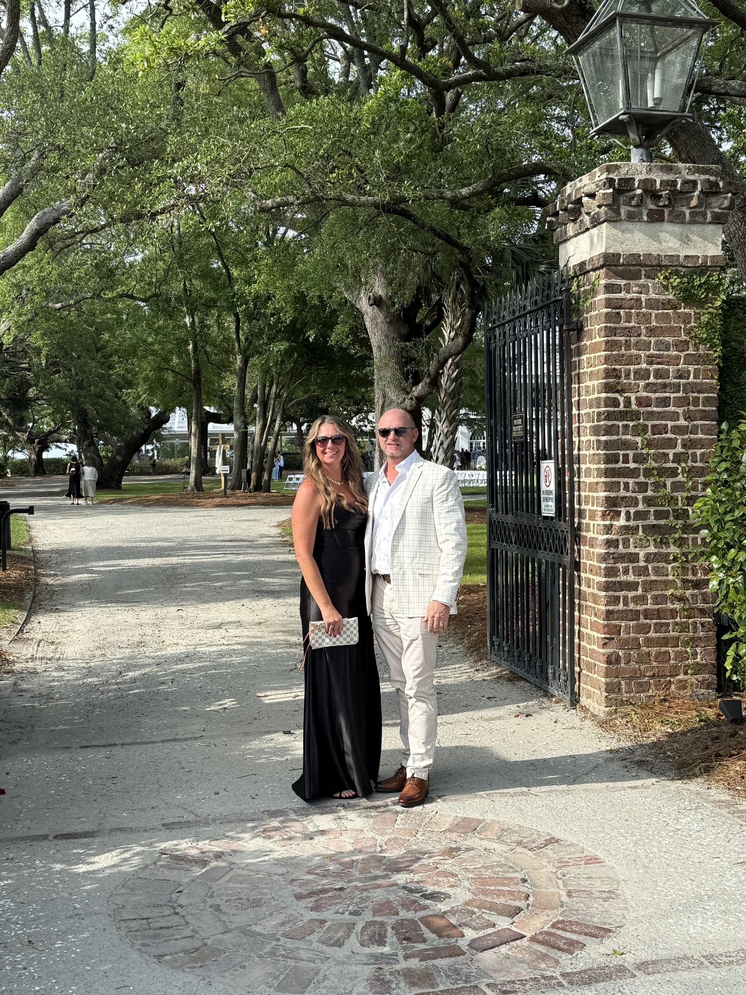 A man and woman dressed in formal attire standing together outdoors in front of a brick gate with a lantern on top. They are smiling and posing for the photo with trees and other people in the background.