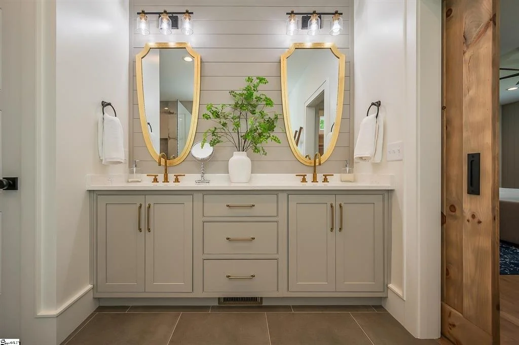 Bathroom vanity with two mirrors, golden fixtures, white countertop, gray cabinet, and a potted plant in the center.
