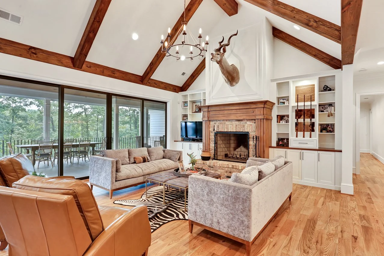Living room with a vaulted ceiling, wooden beams, a mounted antelope head above a brick fireplace, large sliding glass doors opening to an outdoor deck, and cozy seating including a tan leather armchair and light-colored sofas.