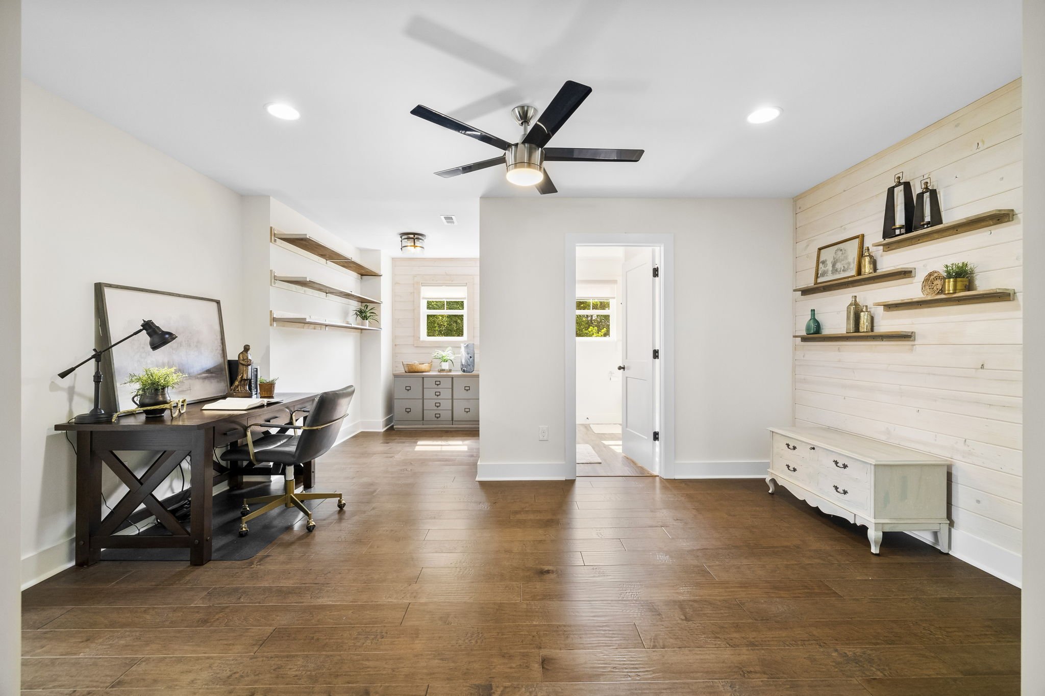 Interior view of a modern living space with white walls, wooden flooring, a black desk with a lamp, decorative items, and shelves with books and decor, illuminated by ceiling lights and natural light from windows.