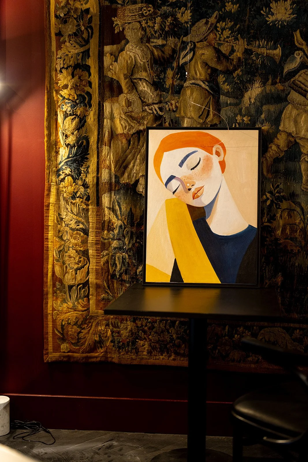 Een schilderij van een vrouw met rood haar en gesloten ogen, haar hoofd rustend op haar schouder, geplaatst op een zwarte tafel. Achter de tafel hangt een grote, getailleerde muurhanger met een historische, rijk versierde textiele wand. Een stoel en een elektrische stekker liggen op de vloer.