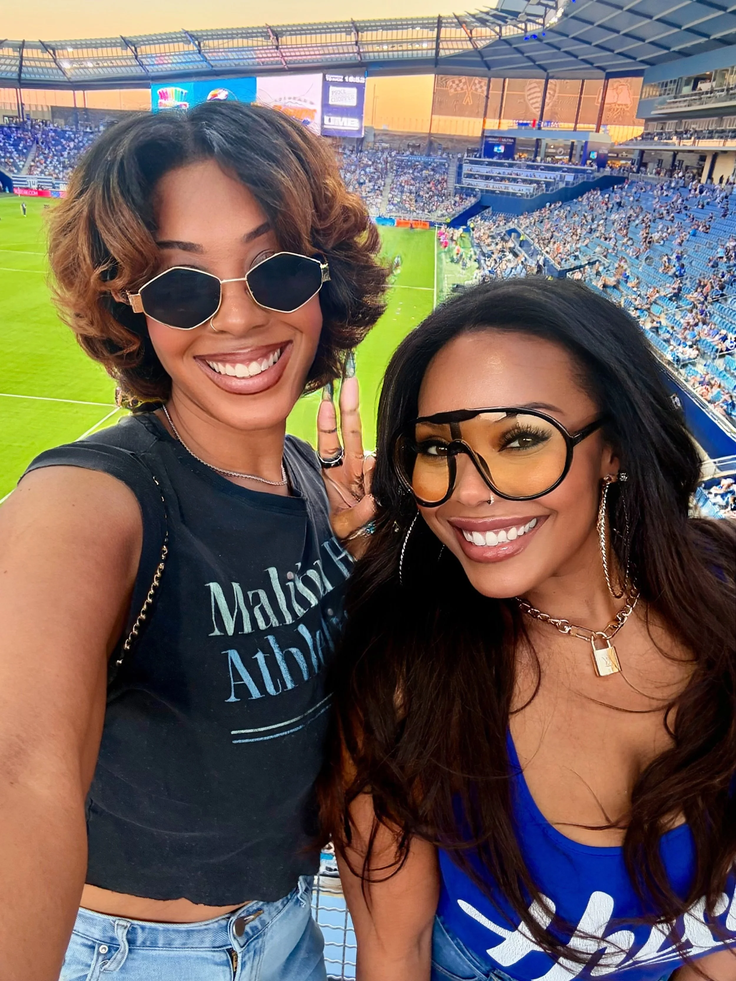 Two women at a sports stadium taking a selfie with a soccer field in the background.