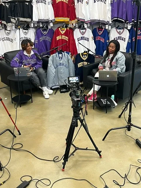 Two women are seated in a recording studio surrounded by sports jerseys hanging on racks, with cameras and lights set up for recording.