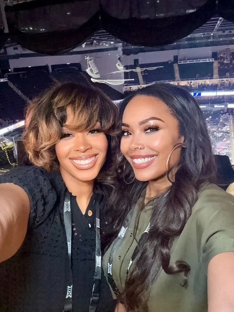 Two women smiling and taking a selfie at an indoor event or concert, with a large stage and seating visible in the background.