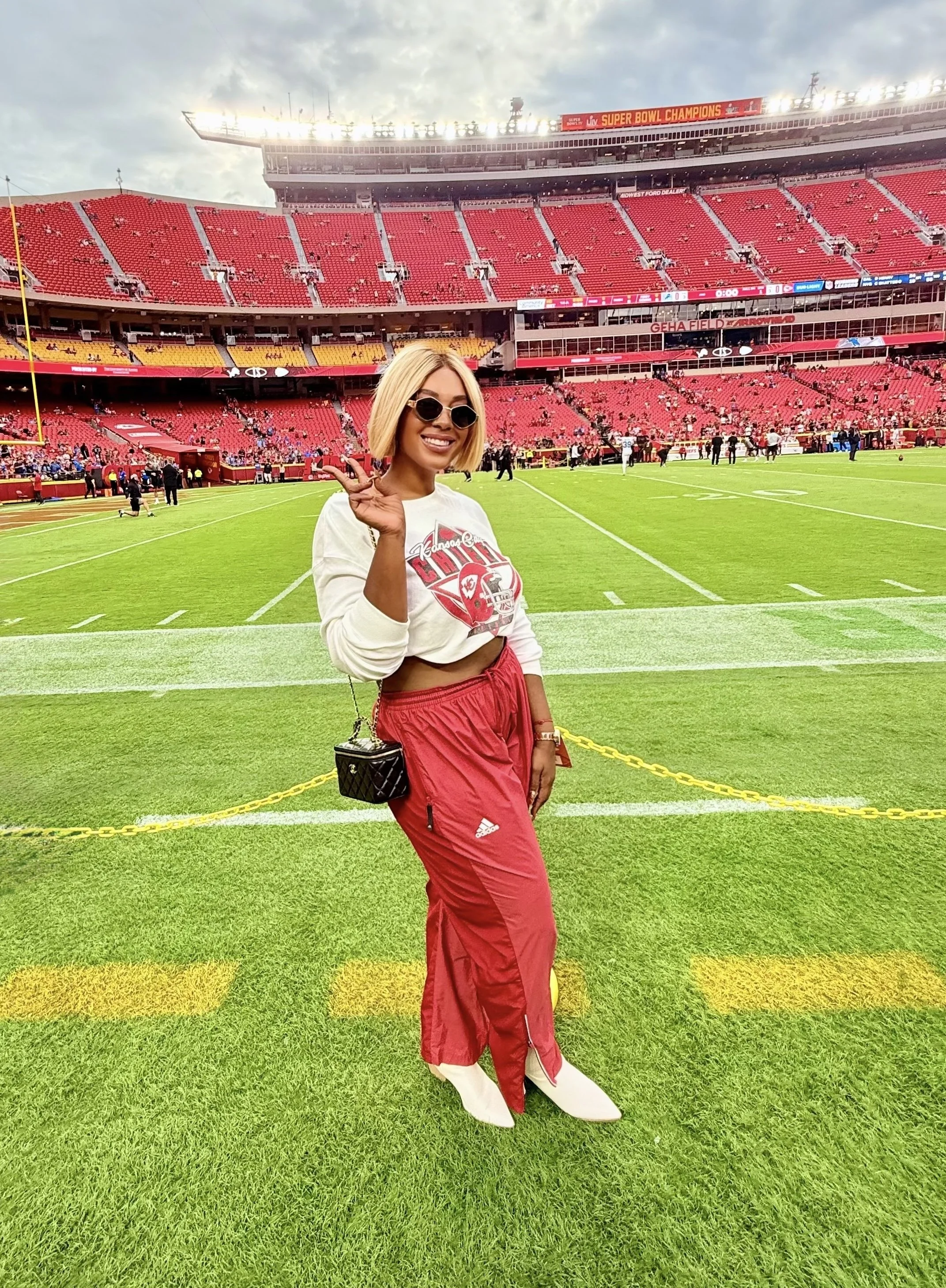 A woman with standing on a football field at a stadium, smiling, wearing sunglasses, a white cropped sweatshirt with a logo, red track pants, white boots, and carrying a small black purse, with the stadium seats and scoreboard in the background.