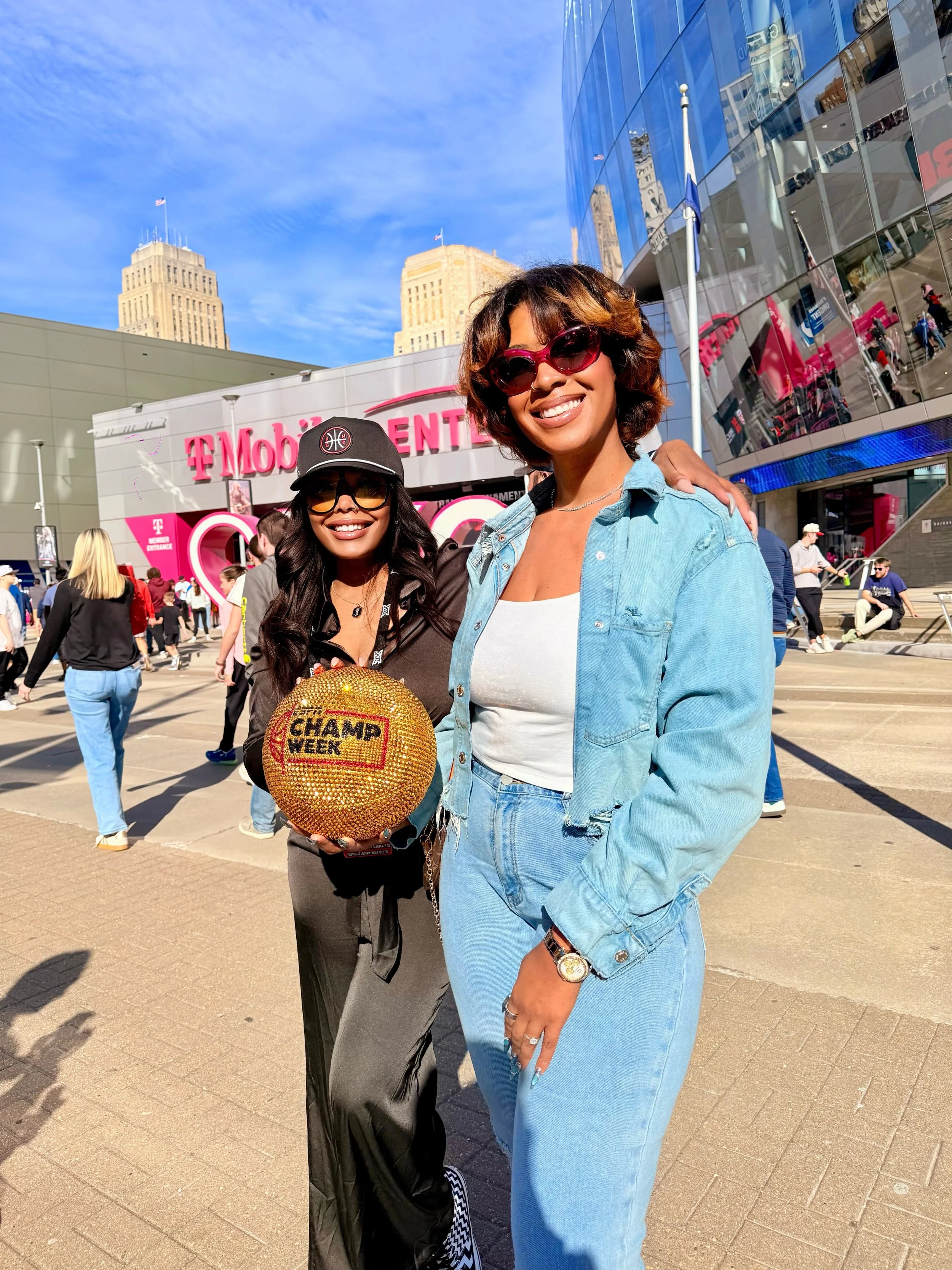 Two women smiling outdoors in front of a T-Mobile store during daytime, one holding a sparkly volleyball with the words 'Champ Week,' people walking in the background, and tall buildings under a blue sky.
