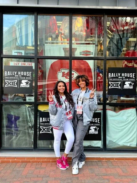 Two women standing in front of a glass window decorated with Kansas City Chiefs and rally house banners, making peace signs.