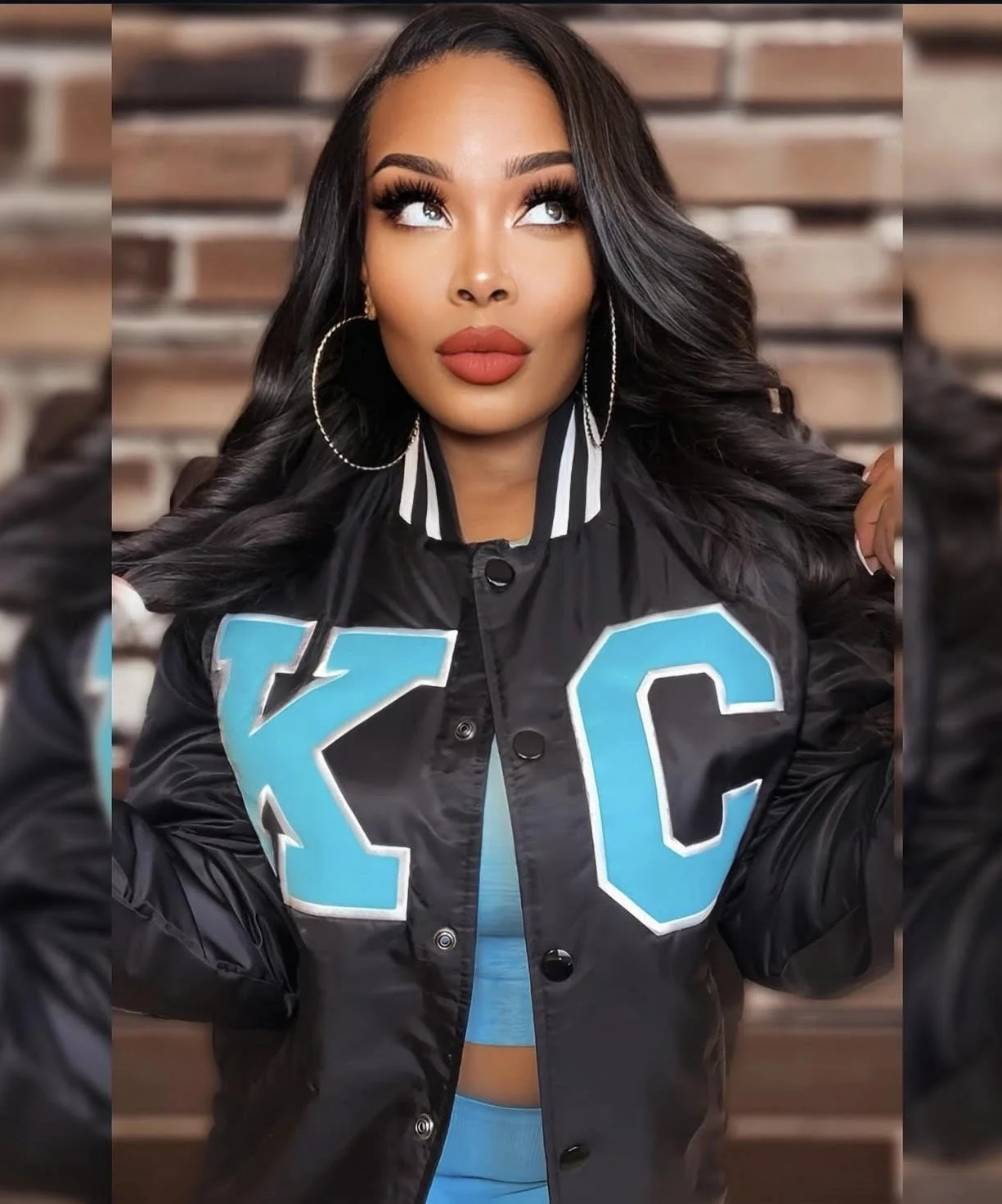 A woman with long, wavy black hair, large gold hoop earrings, and bold makeup including red lipstick and eye makeup, wearing a black bomber jacket with blue Greek letters and a blue top underneath, standing in front of a wooden background.