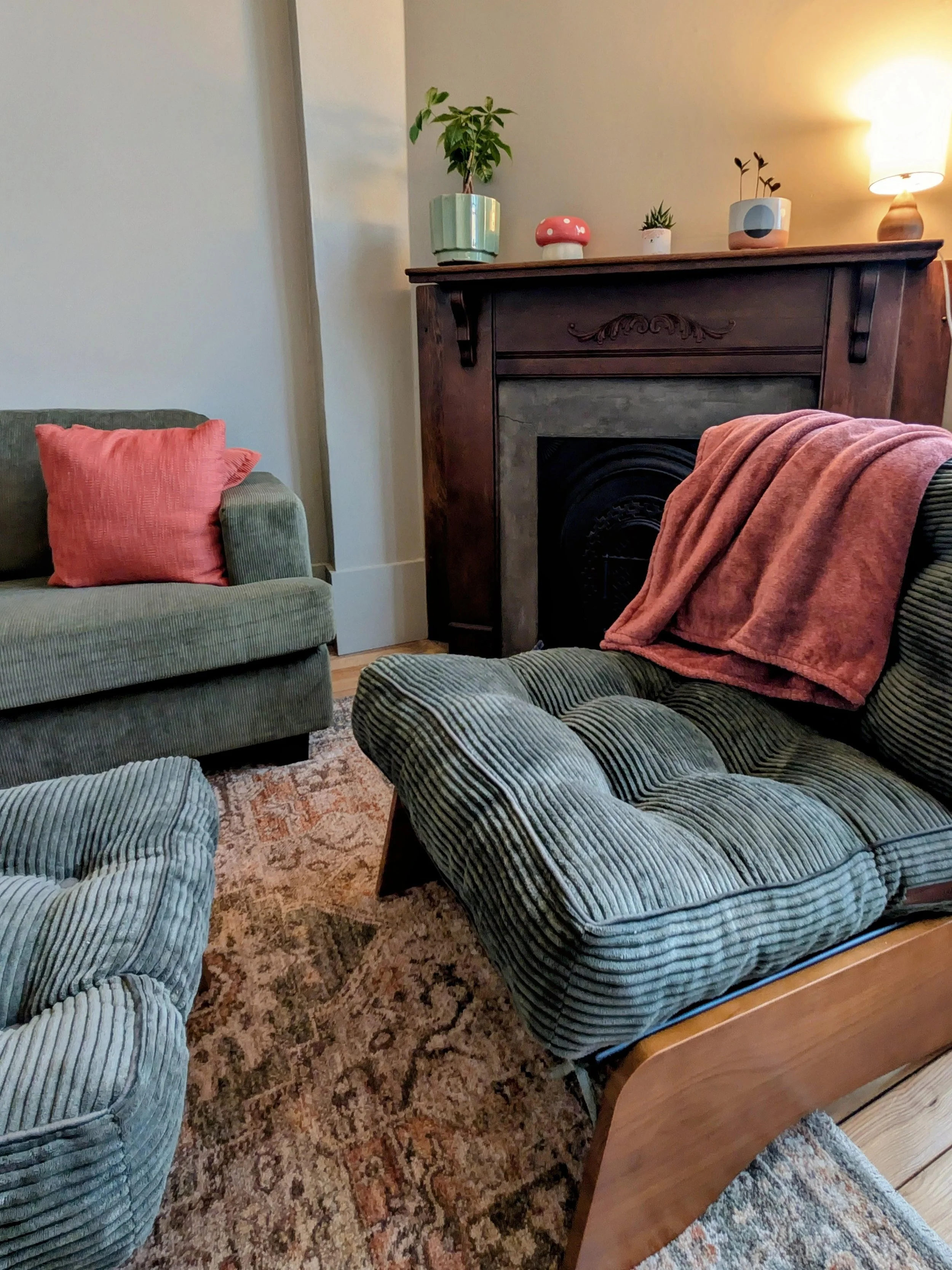 Living room with a green armchair, a fireplace mantel decorated with potted plants and a mushroom-shaped decor, a lamp in the background, and a wooden side table with a pink towel.