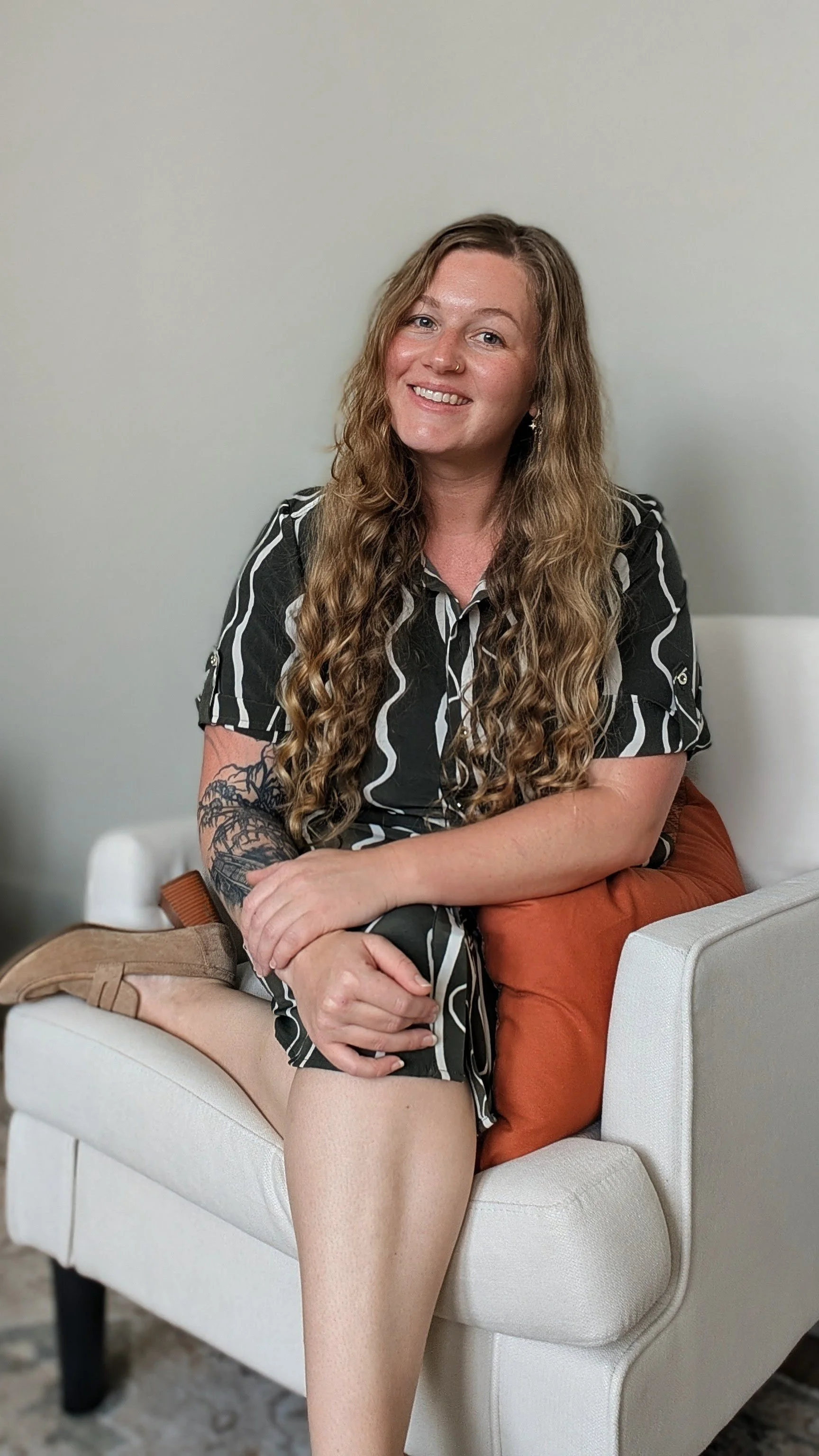 A woman with long wavy hair, wearing a black and white striped dress, sitting on a white couch with a brown pillow, smiling at the camera.