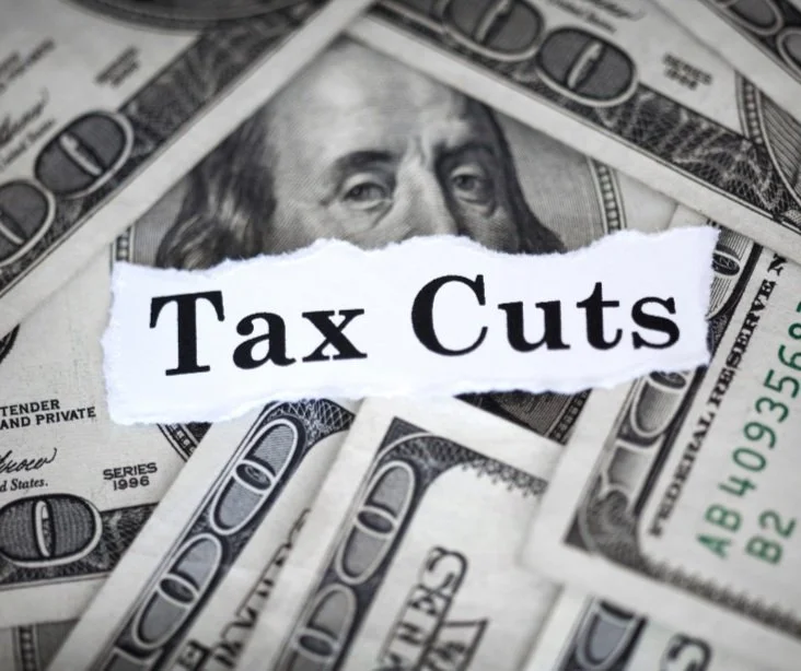 A torn paper with the words "Tax Cuts" placed over a pile of US hundred-dollar bills.