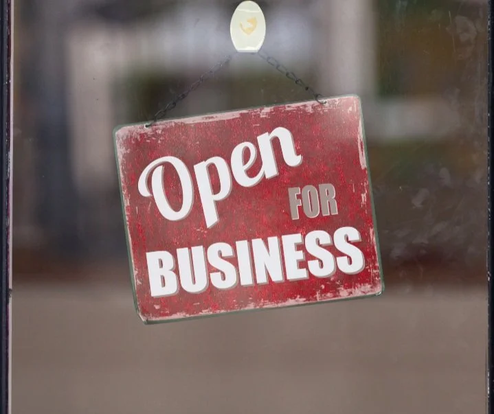 A red, weathered sign hanging from a chain on a glass door reads 'Open for Business.'
