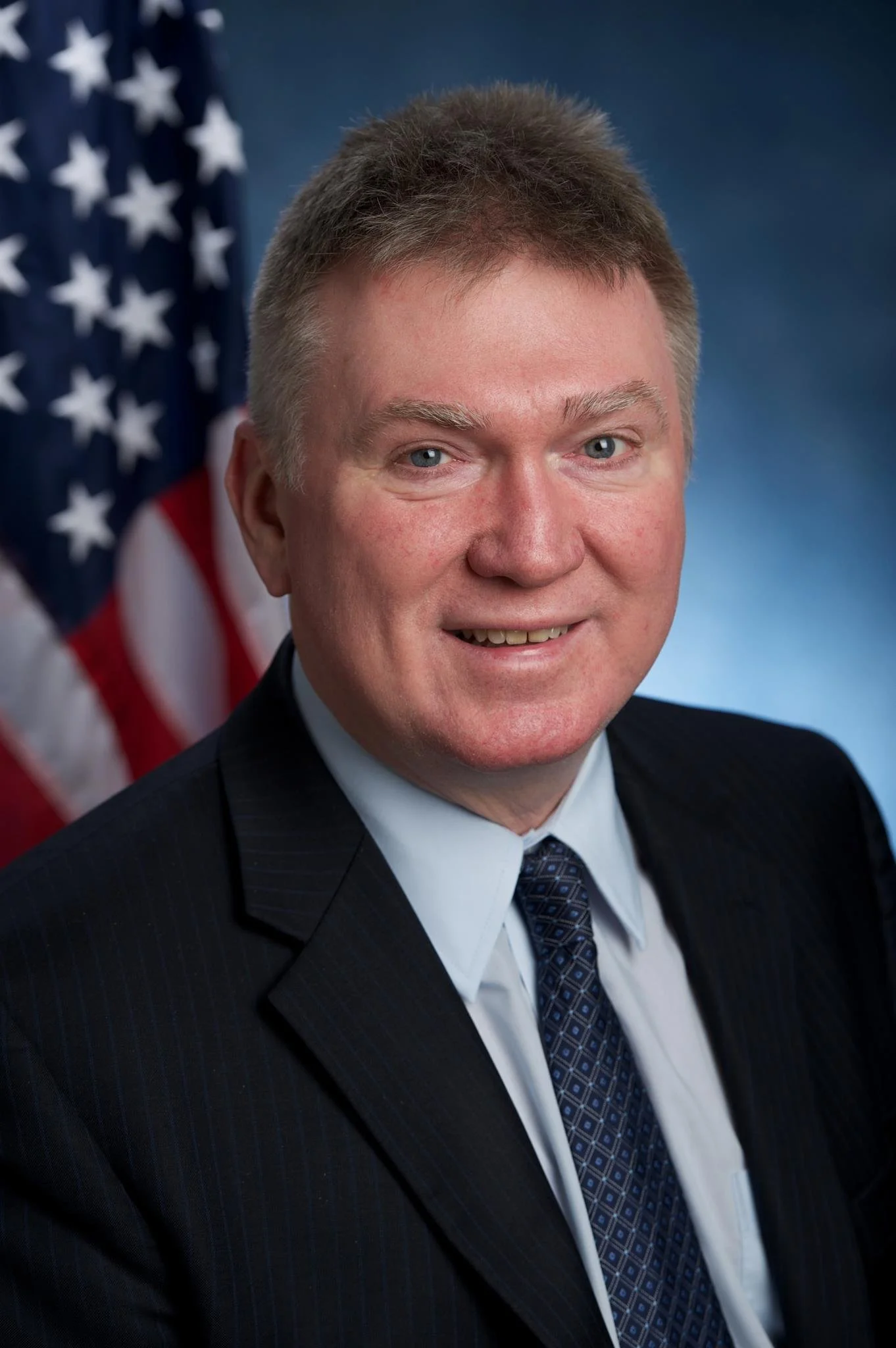 Portrait of a middle-aged man with short light brown hair, wearing a dark pinstripe suit, white shirt, and a patterned tie, standing in front of an American flag and a blue background.