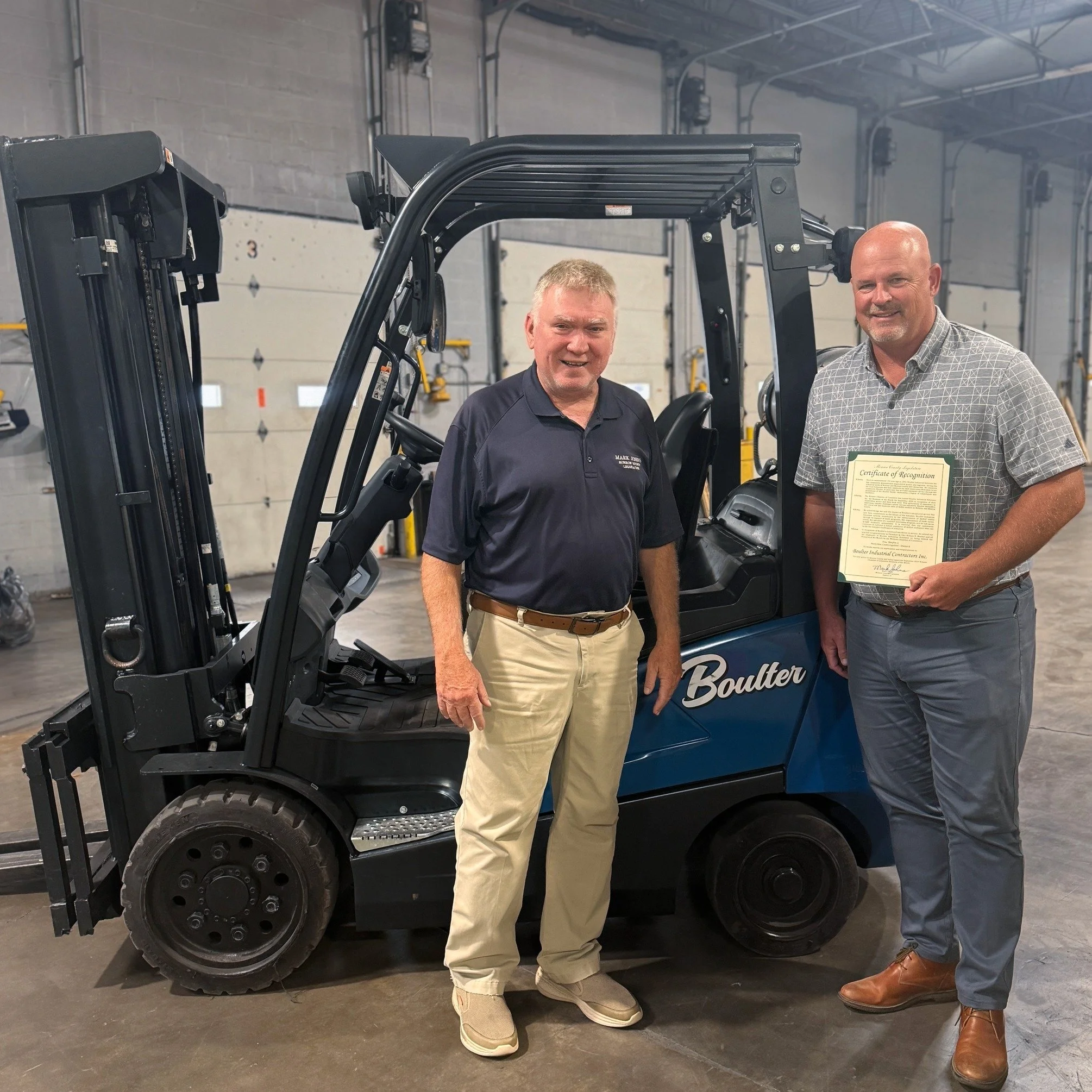 Two men standing inside a warehouse next to a blue forklift, one holding a certificate of recognition.