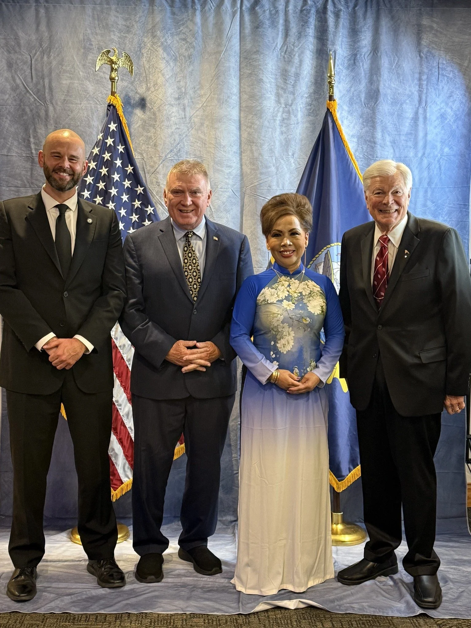 Four people standing in front of American and state flags, dressed in formal attire, smiling for the photo.