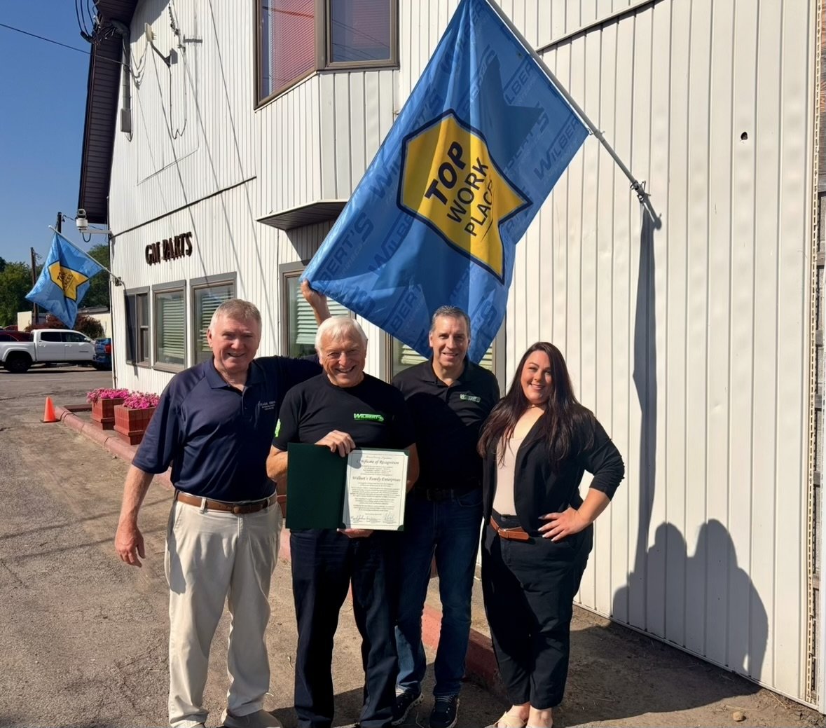 Four people standing in front of a building with flags, one holding a framed certificate, smiling at the camera, sunny day.