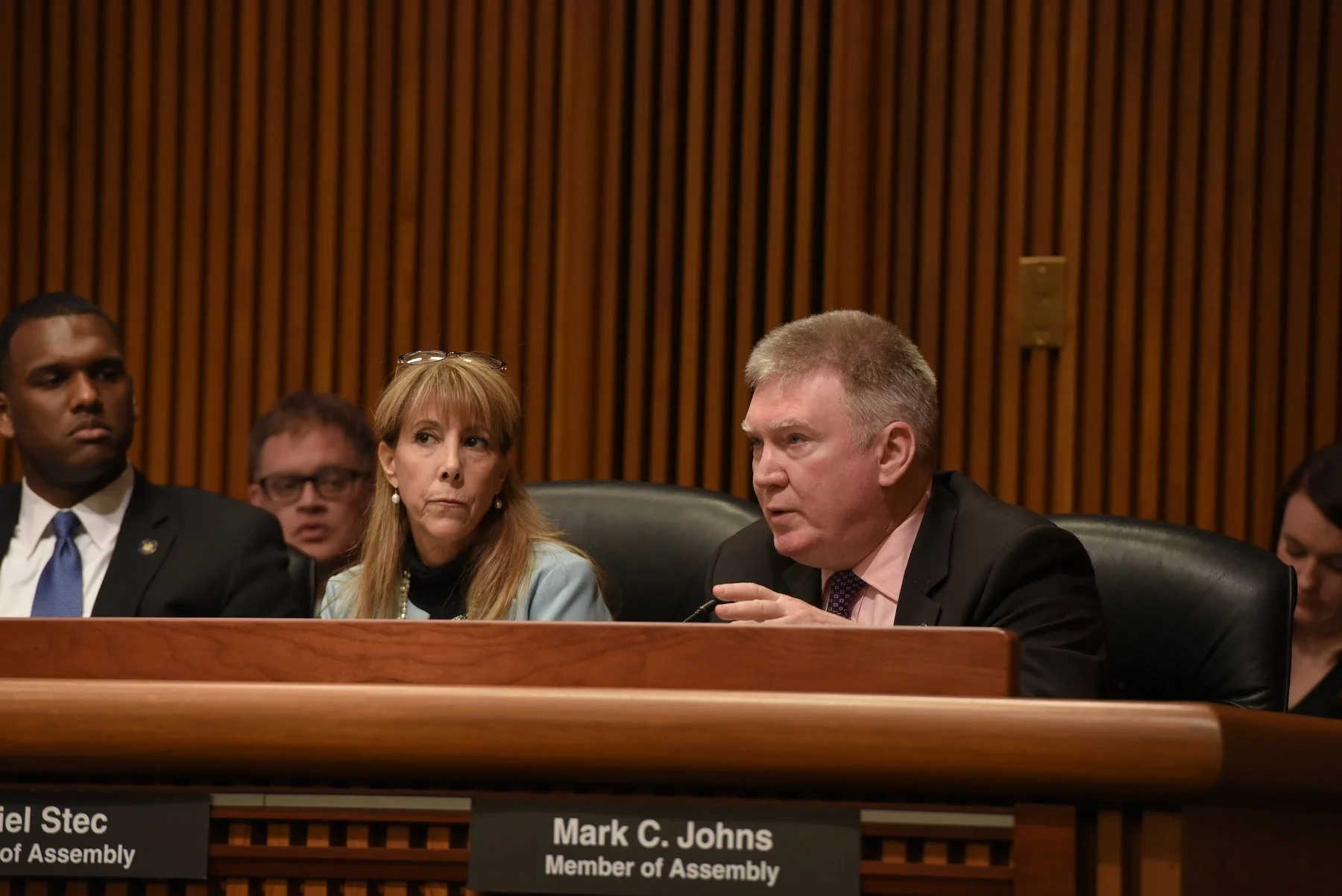 A group of people sitting at a panel discussion. The man on the right is speaking, with a nameplate reading Mark C. Johns, Member of Assembly.