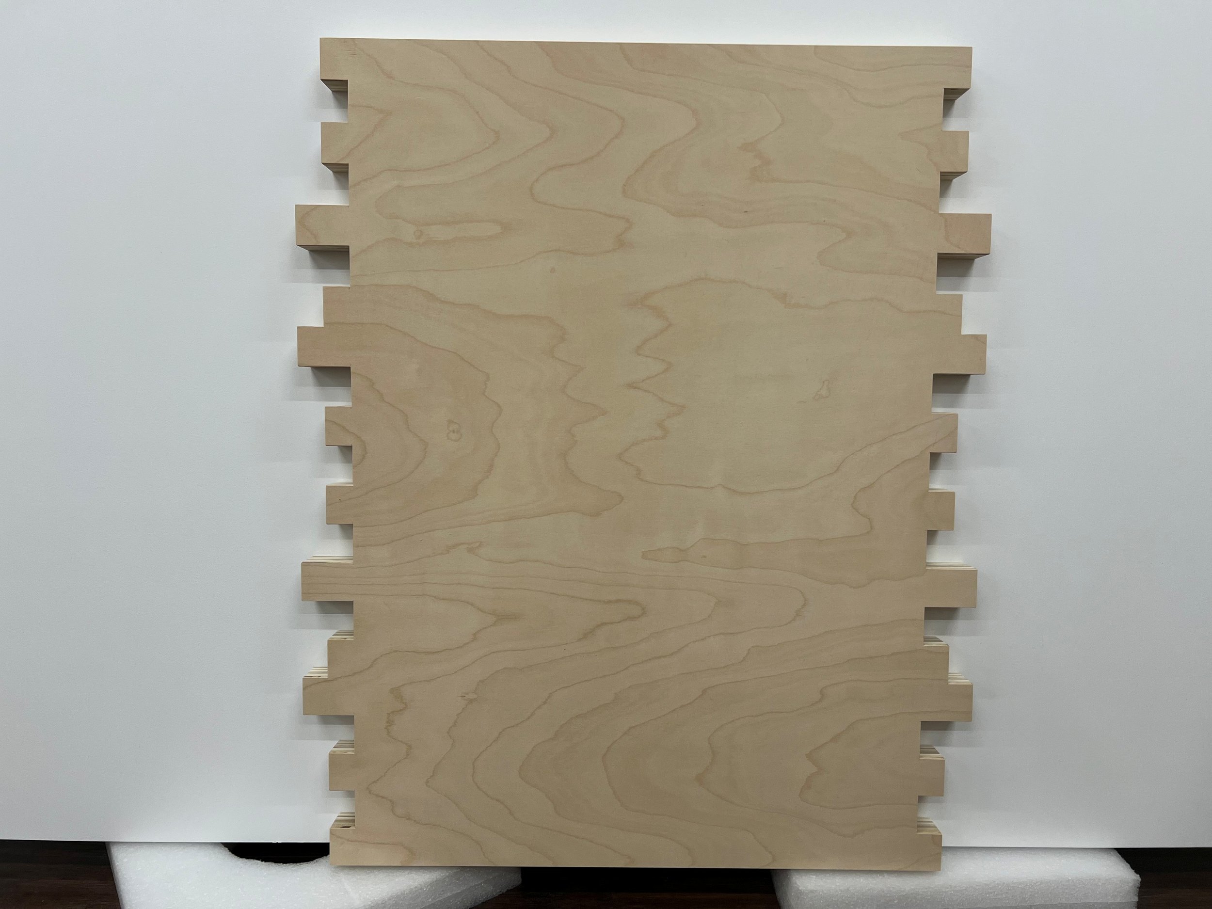 A piece of light-colored wood with rectangular notches on the left side, placed on white foam padding against a white wall.