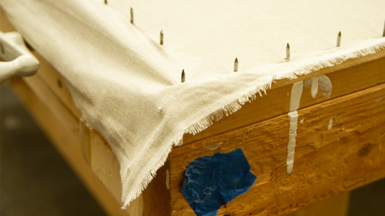 Close-up of a wooden furniture rack of canvas stretched and tightened on a rack, and a small blue patch of tape on the wood.