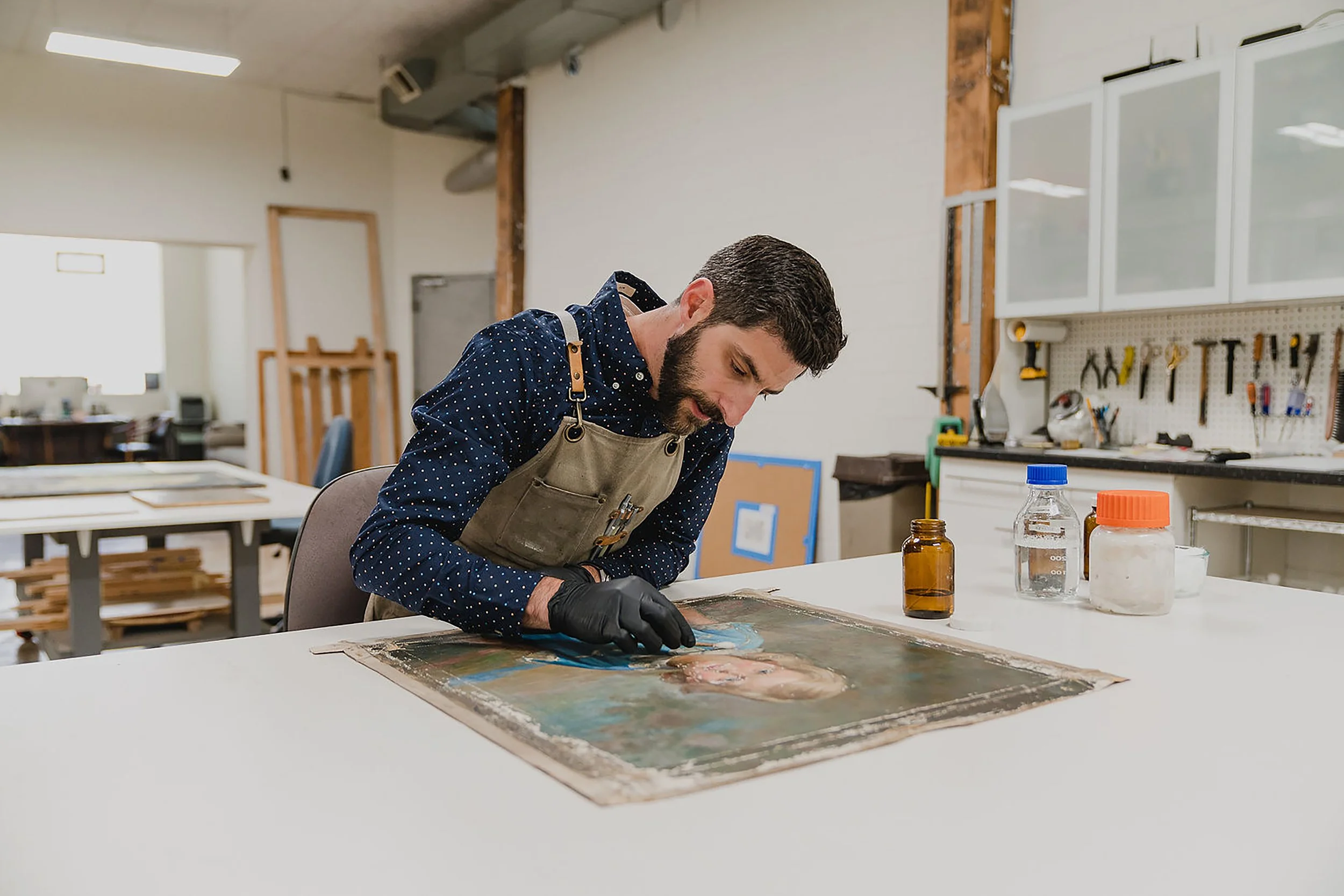 A man with dark hair, beard, wearing a polka dot shirt, beige apron, and black gloves, is working on an art project at a white table in a workshop, with various art supplies around him.