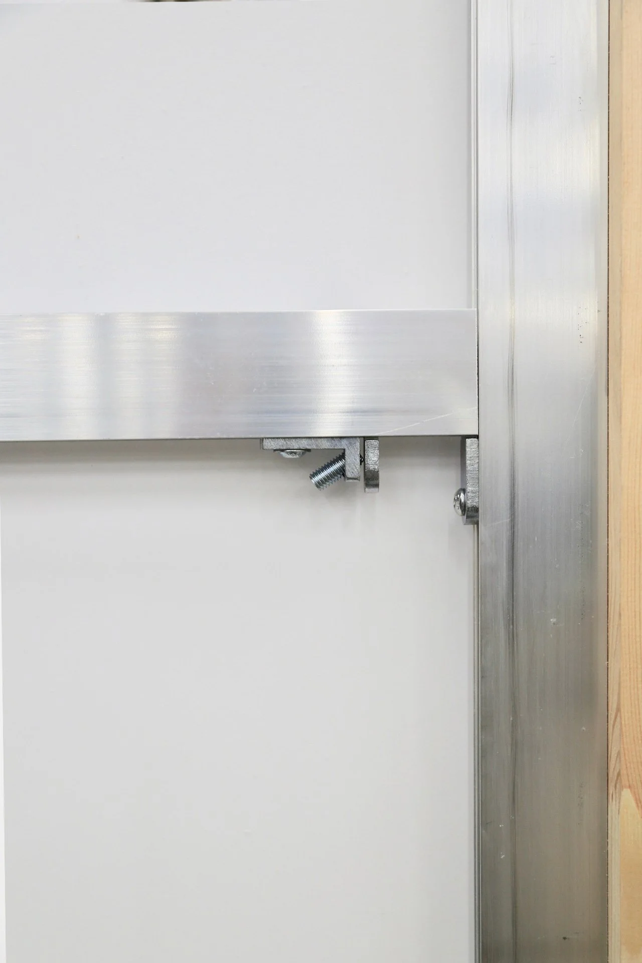 Close-up of a metal hinge and bracket attached to a white wall panel and a vertical metal frame with a wooden edge.