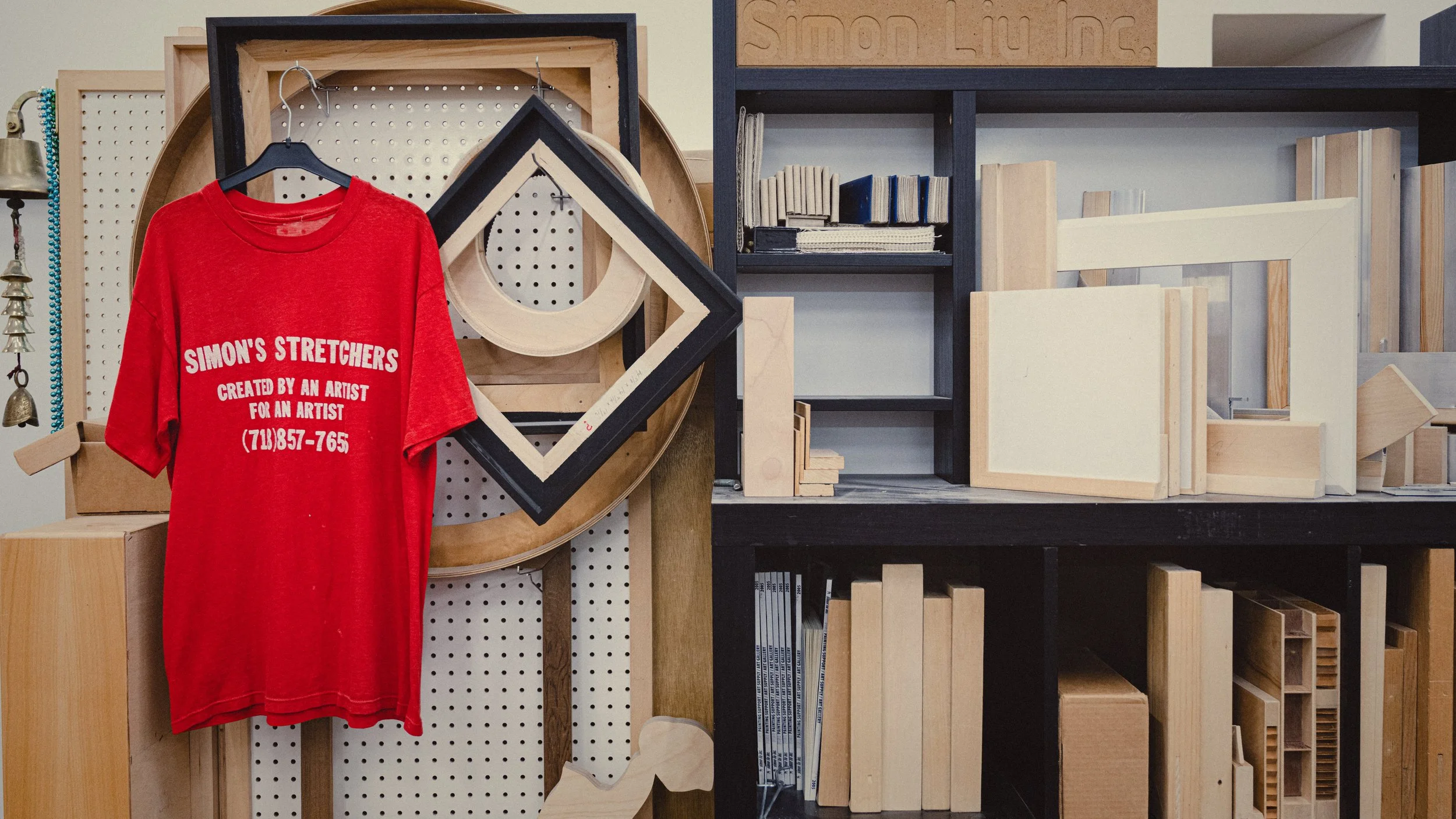 Vintage Simon Liu, Inc. red T-shirt hanging on a black hanger in front of samples of our art support products in the background.