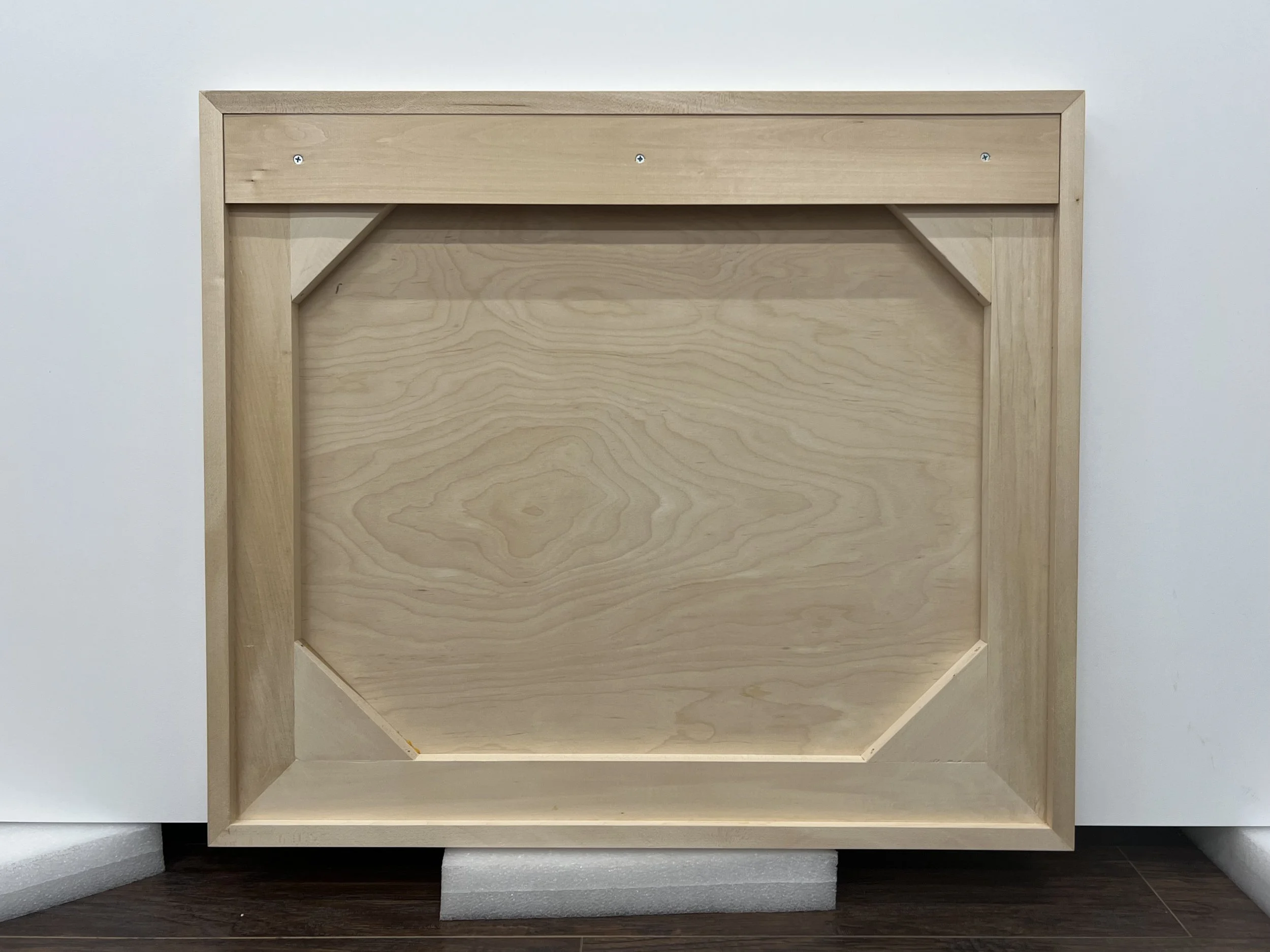 Unfinished wooden picture frame with a light wood finish, placed on a white foam block against a white wall.
