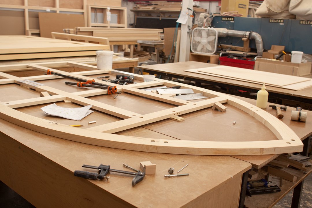 A woodworking workshop with a large table holding an unfinished wooden frame, clamps, glue, and other tools. Background includes shelves, a fan, and woodworking machinery.