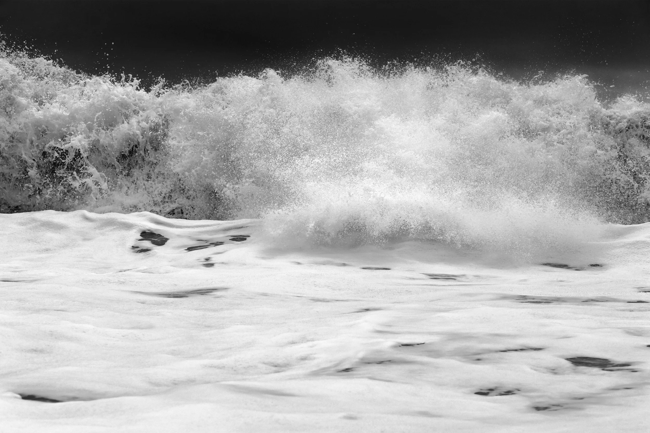 Black and white photograph of ocean waves crashing onto a shoreline with foam and spray.