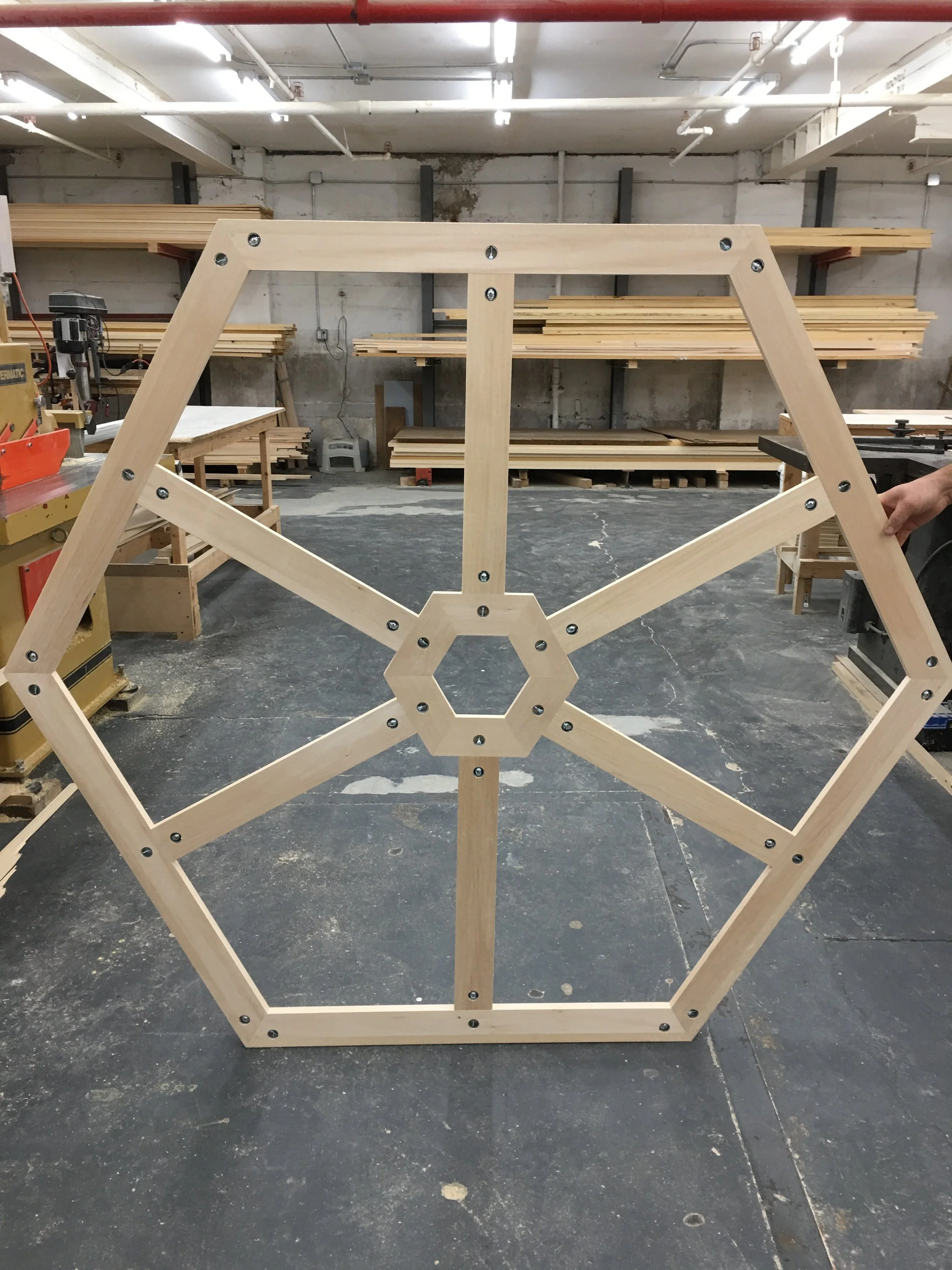 A wooden geometric structure with hexagon and triangle shapes held together with screws, situated in a woodworking workshop with shelves of wood planks in the background.