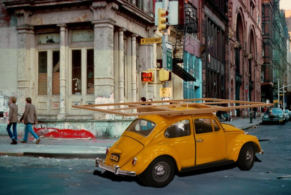 A yellow vintage Volkswagen Beetle parked on a city street with a long wooden art support (stretcher) on top. The background shows old Soho (New York City), a street corner with a stop sign, and pedestrians walking on the sidewalk.