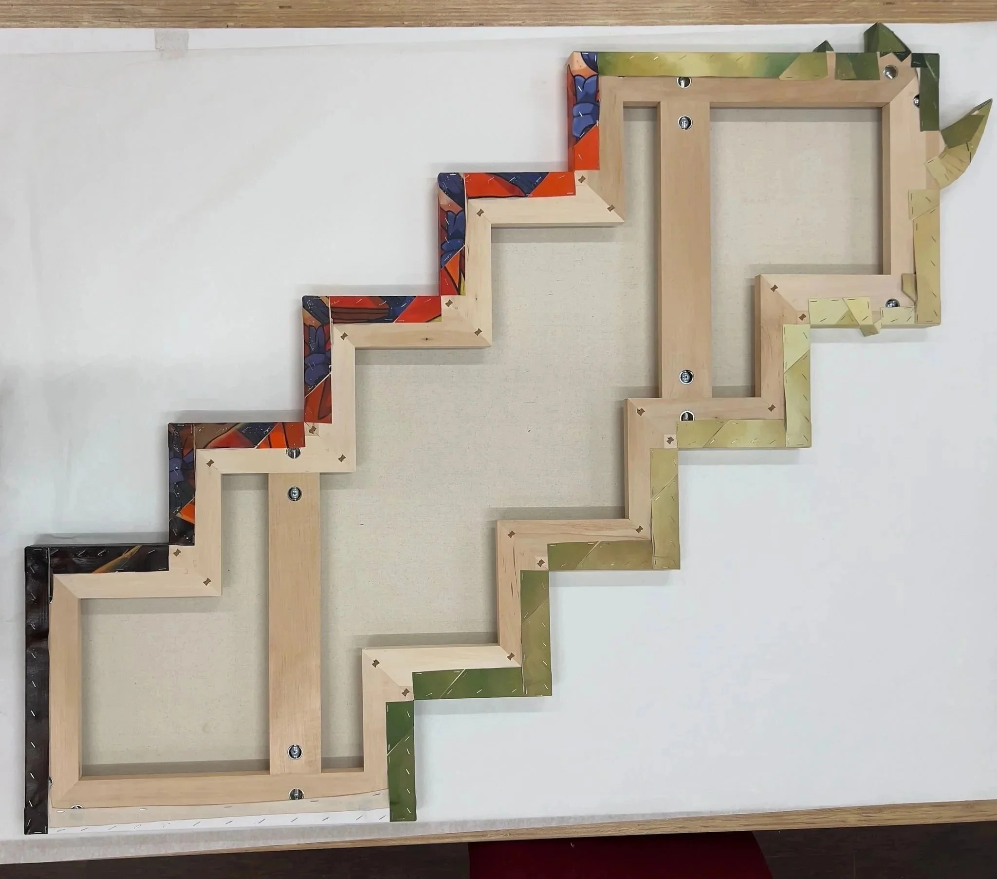Unassembled wooden picture frame with colored decorative panels at the corners, screws, and corner braces, mounted on a plain white wall.