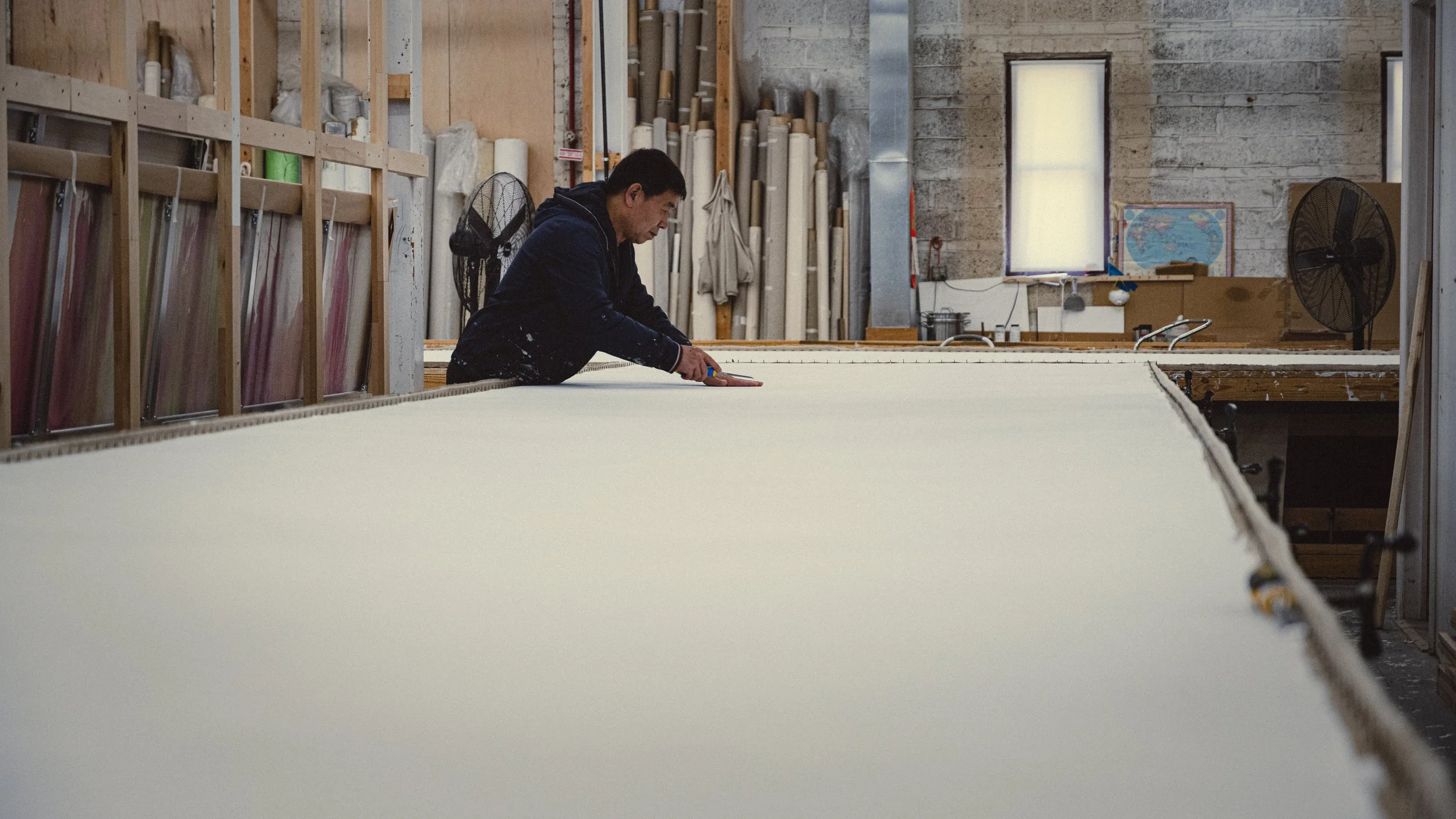 A man working on a large piece of material in a workshop with shelves of supplies behind him.