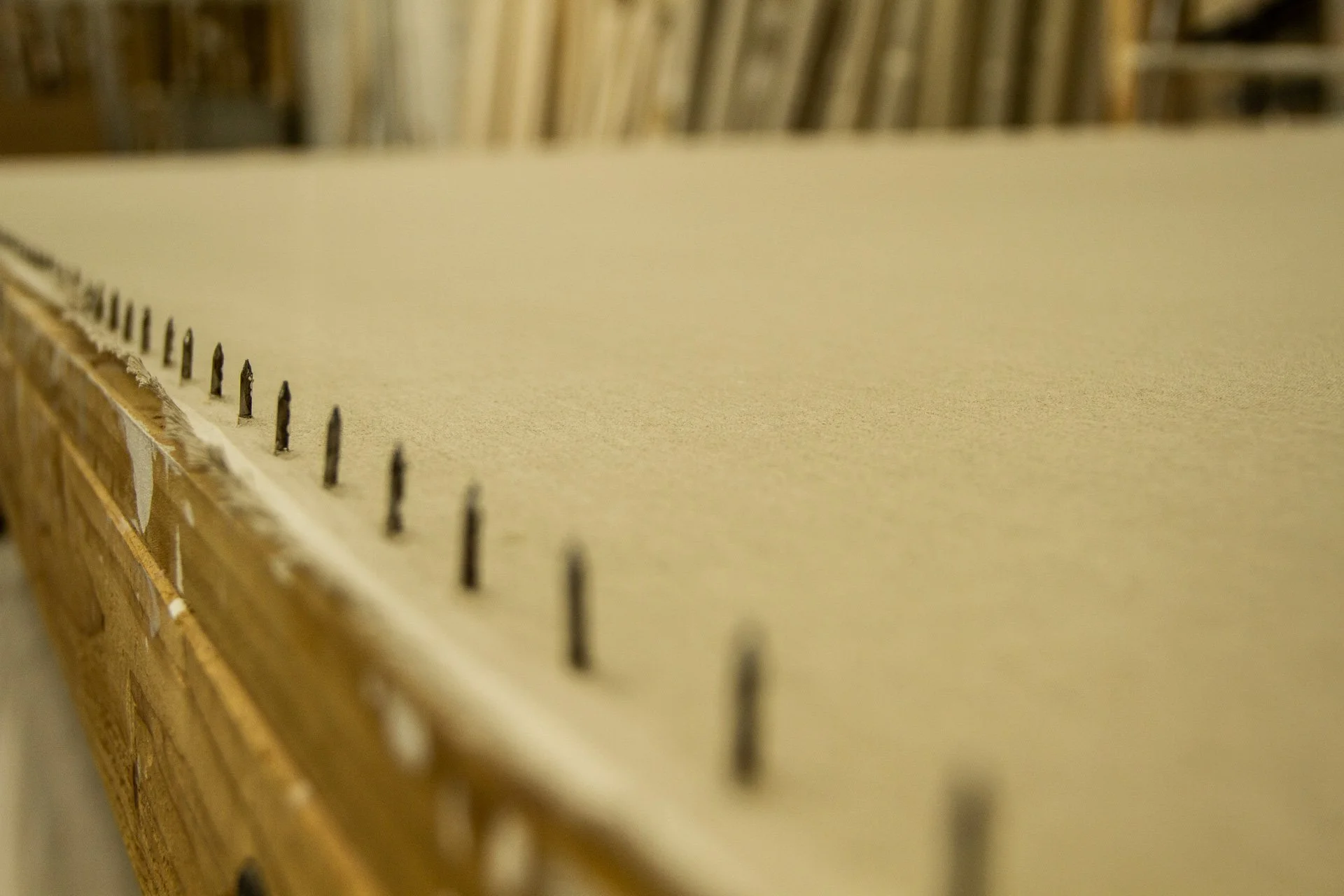 Close-up view of a large sheet of beige drywall with several metal nails lined up along the edge, with a background of stacked drywall sheets and wooden pallets.