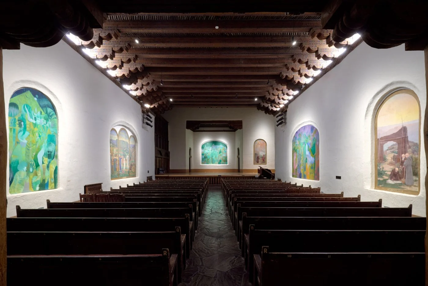 Interior of a church with colorful paintings on the walls, featuring stretchers measured to the exact specifications of the inset archways, with nature and landscape scenes.