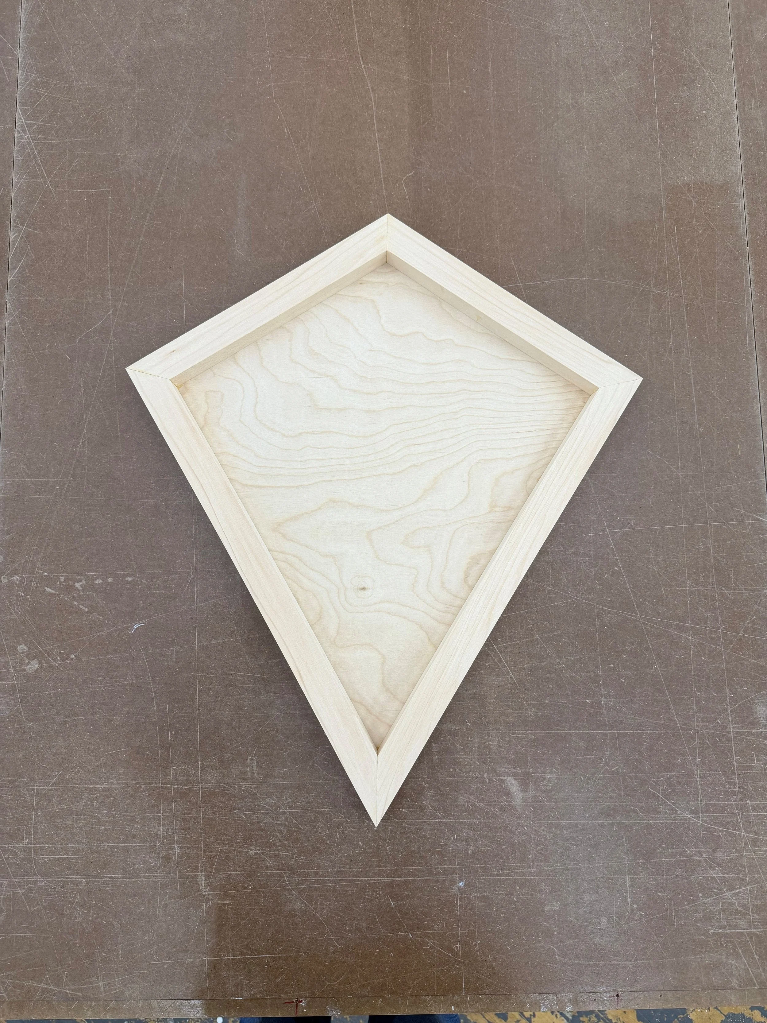 A kite-shaped wooden fine art panel on a brown worktable.