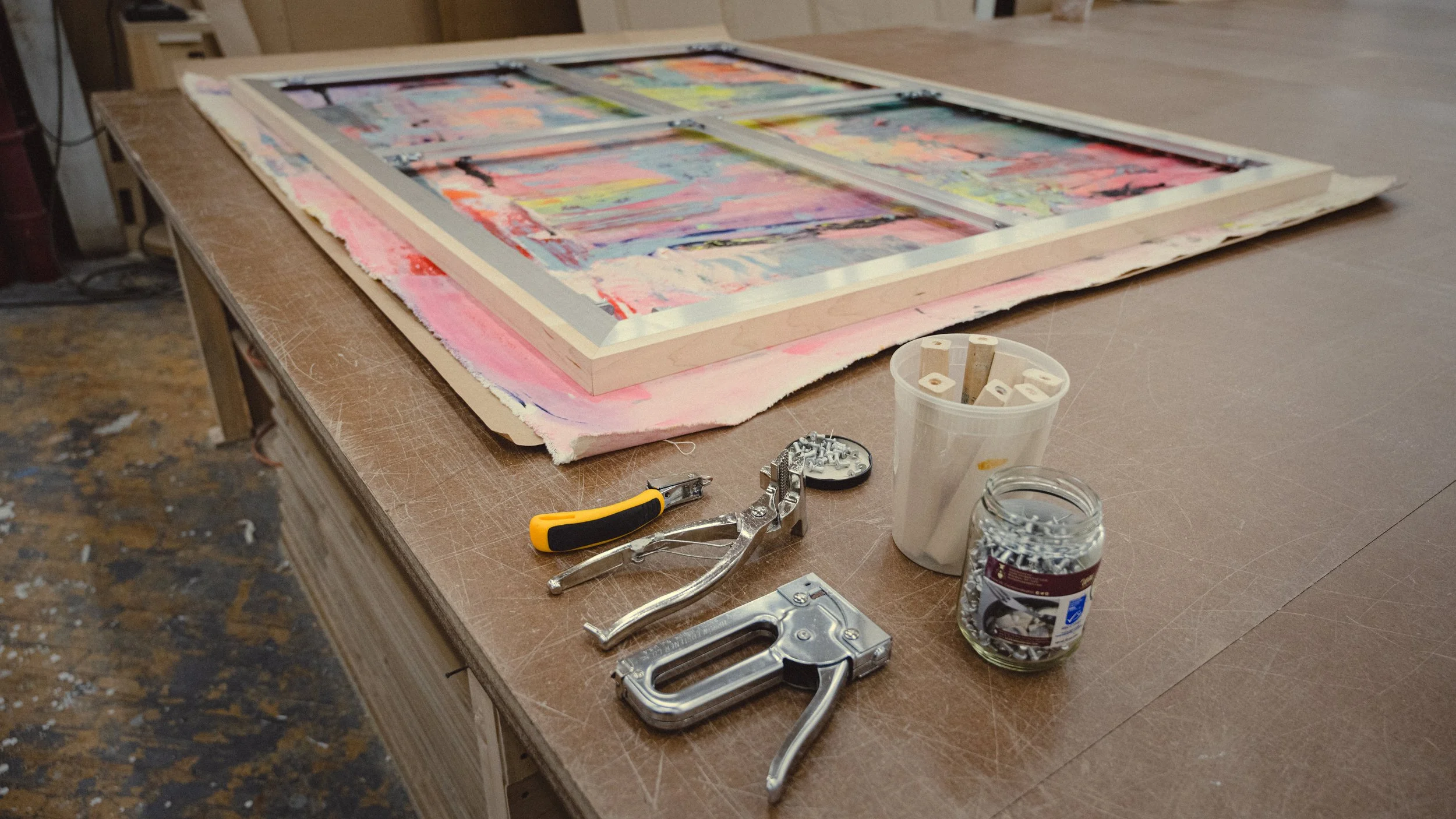 An art studio workspace with a large wooden table containing a colorful abstract painting, tools like staple guns, pliers, a utility knife, jars of nails and screws, and a cup with wooden sticks, indicating an ongoing stretching process.