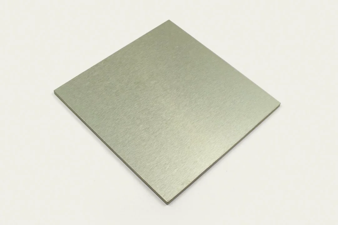 A metallic square chip or plate with a smooth, brushed surface, resting on a light background.