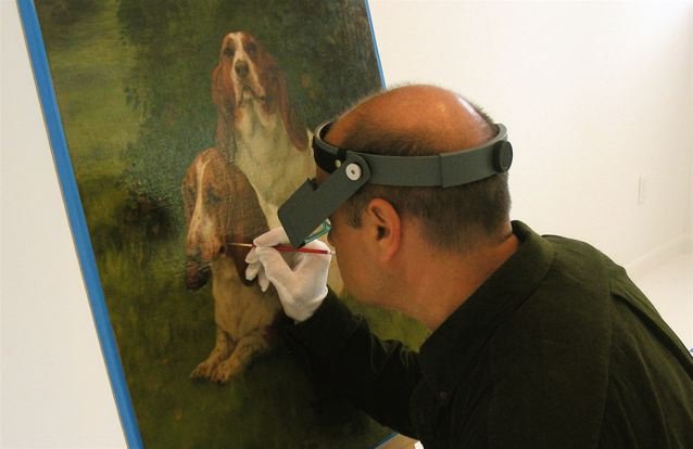 A man wearing a magnifying visor and gloves is carefully restoring or examining a painting of two dogs, one sitting and one lying down.