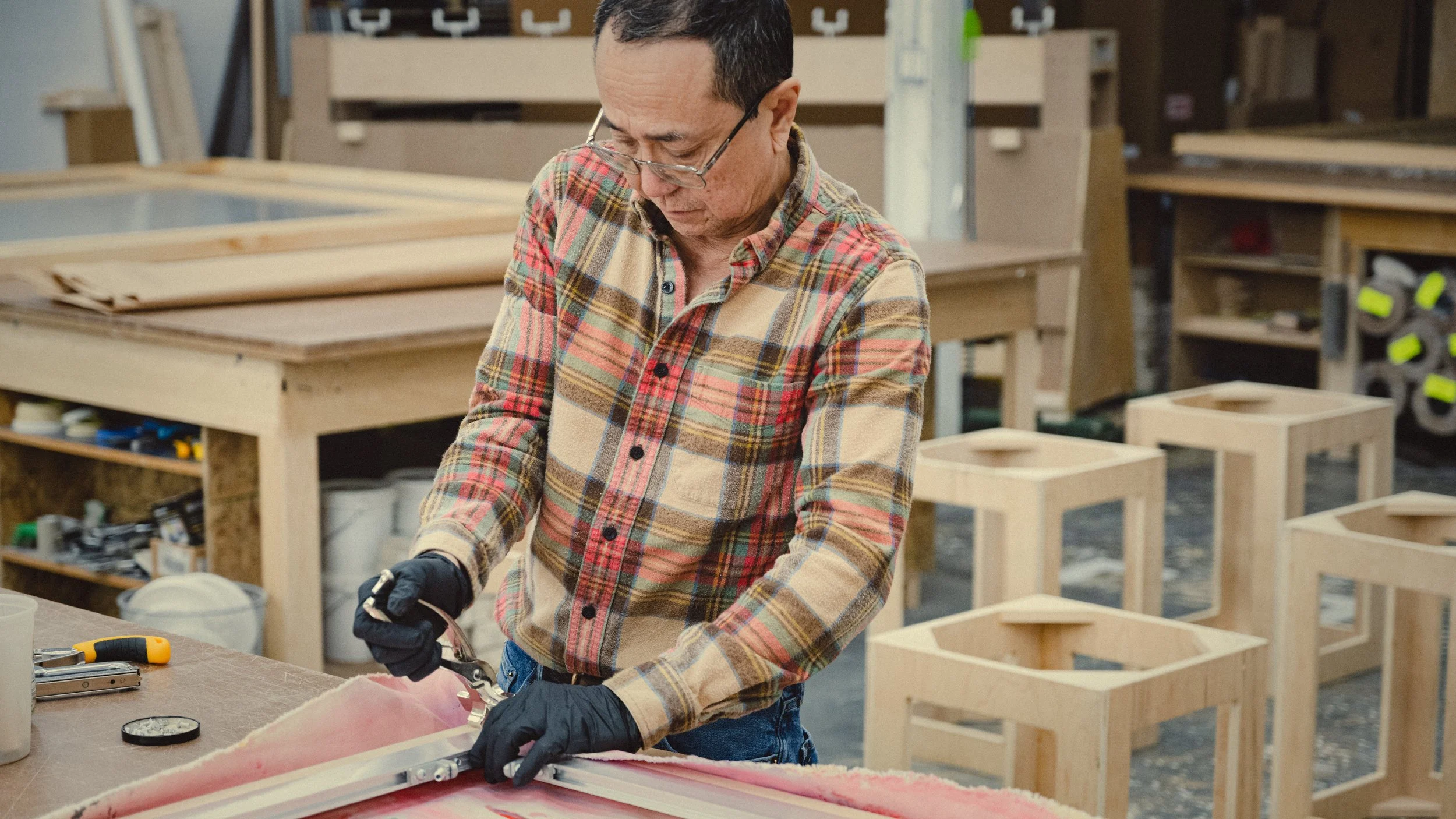 Simon Liu wearing a plaid shirt and glasses working with art stretching tools on a wooden and aluminum stretcher in a woodworking shop with art supports and tools in the background.