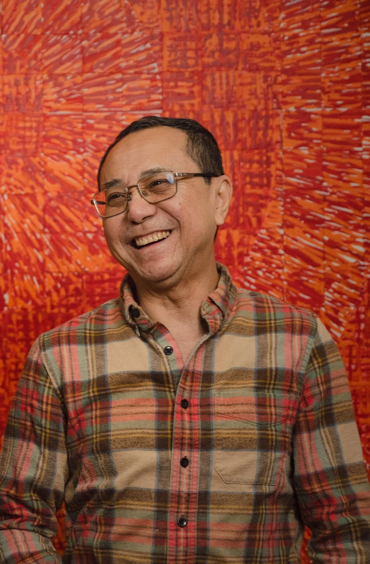 A smiling Simon Liu wearing glasses and a plaid shirt standing in front of his colorful abstract work with red and orange brush strokes.